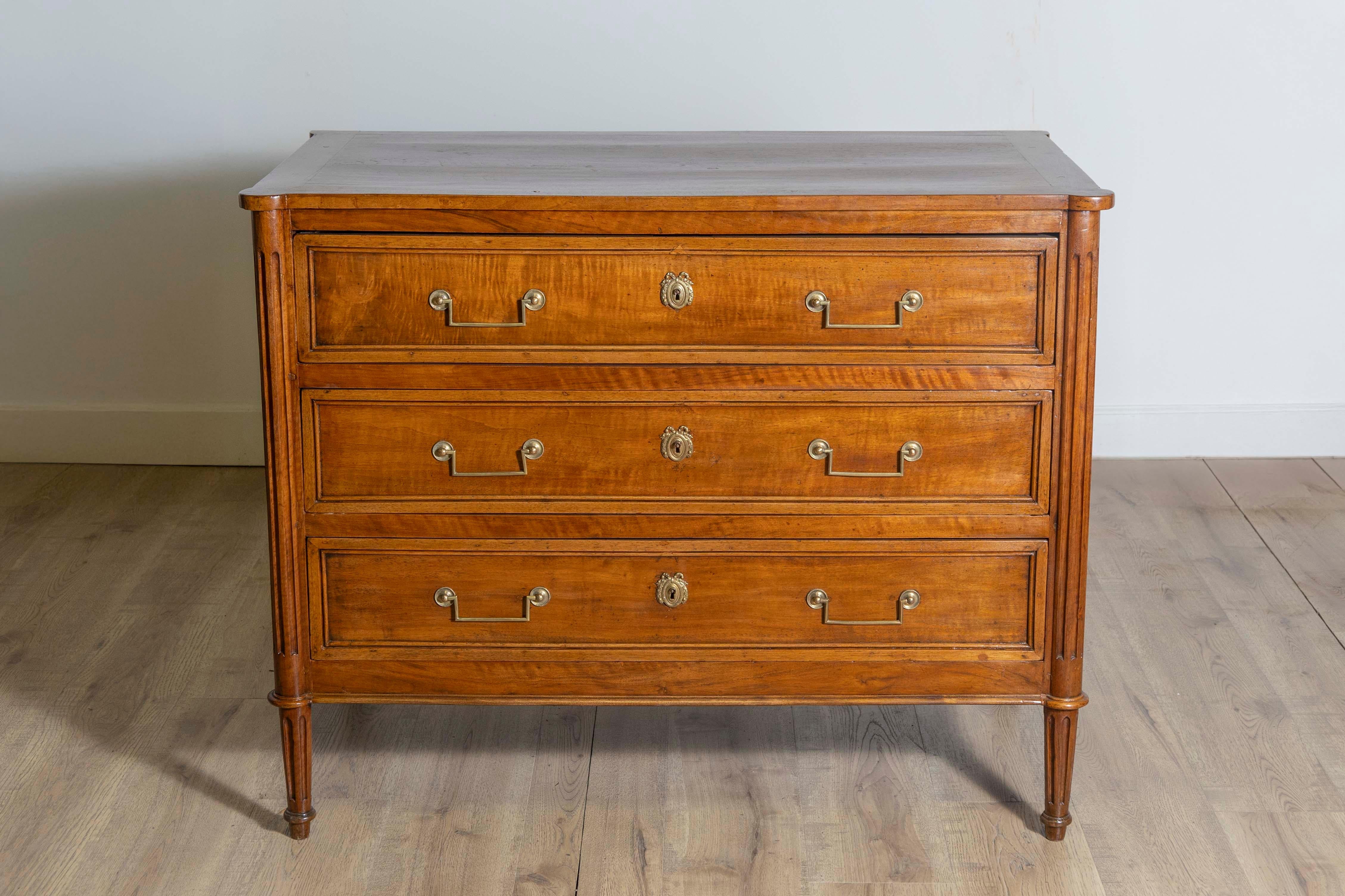 A French Louis XVI period walnut commode circa 1790 with three drawers, linear brass hardware, cylindrical tapered legs and rounded fluted side posts. Step into the elegance of the French Louis XVI period with this exquisite walnut commode from the