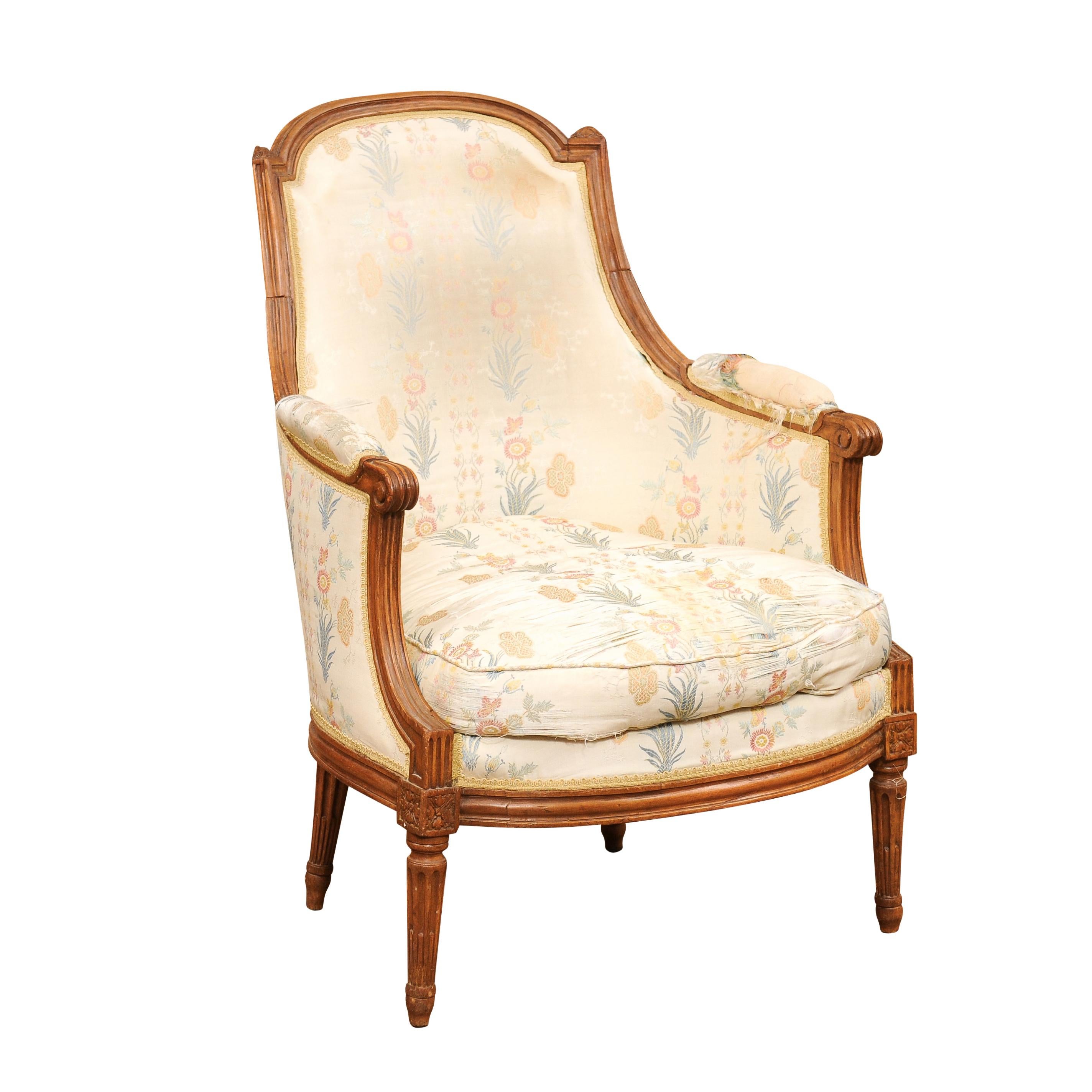 A French Louis XVI period walnut bergère chair from the late 18th century with curving back, scrolling arms, carved rosettes and fluted tapering legs. Experience the timeless elegance of this French Louis XVI period walnut bergère chair from the