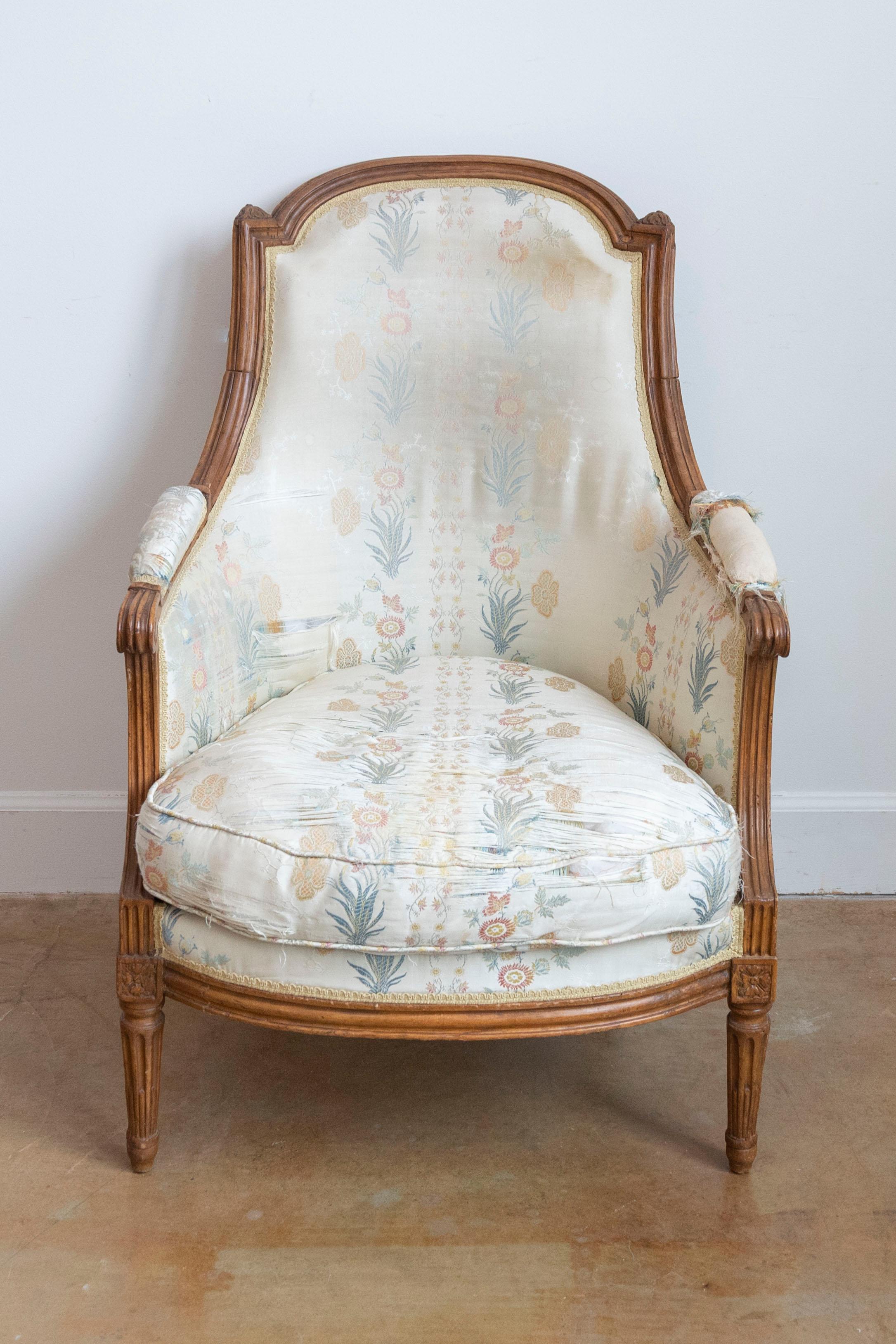 A French Louis XVI period walnut bergère chair from the late 18th century with curving back, scrolling arms, carved rosettes and fluted tapering legs. Experience the timeless elegance of this French Louis XVI period walnut bergère chair from the