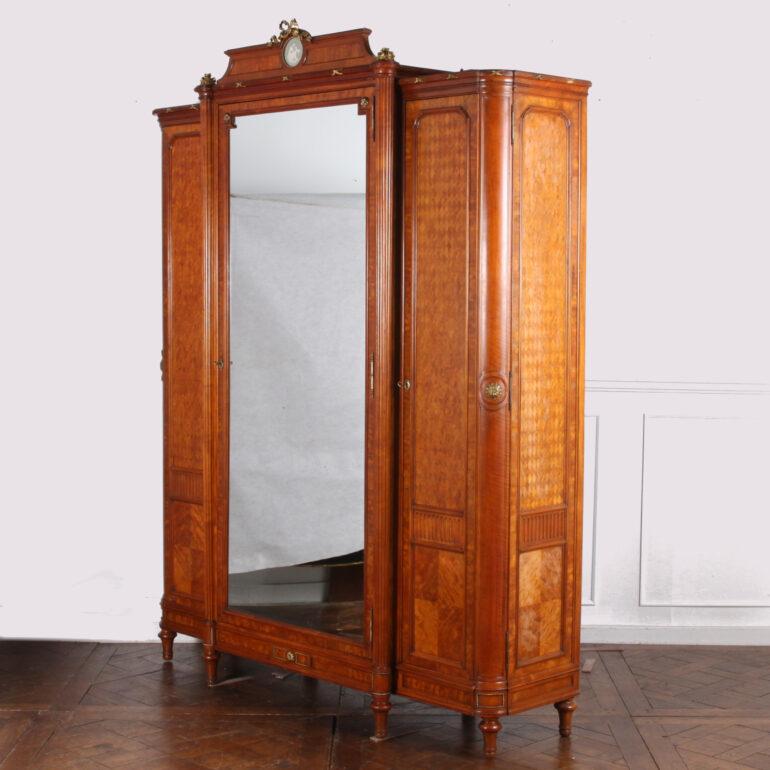 A French Louis XVI style three door armoire in satinwood with parquetry panels and carved details. Fine quality gilt bronze (also known as 'ormolu') mounts and porcelain ‘cameo’ plaque to top crown. Fitted shelves to the interior. Interior mounted