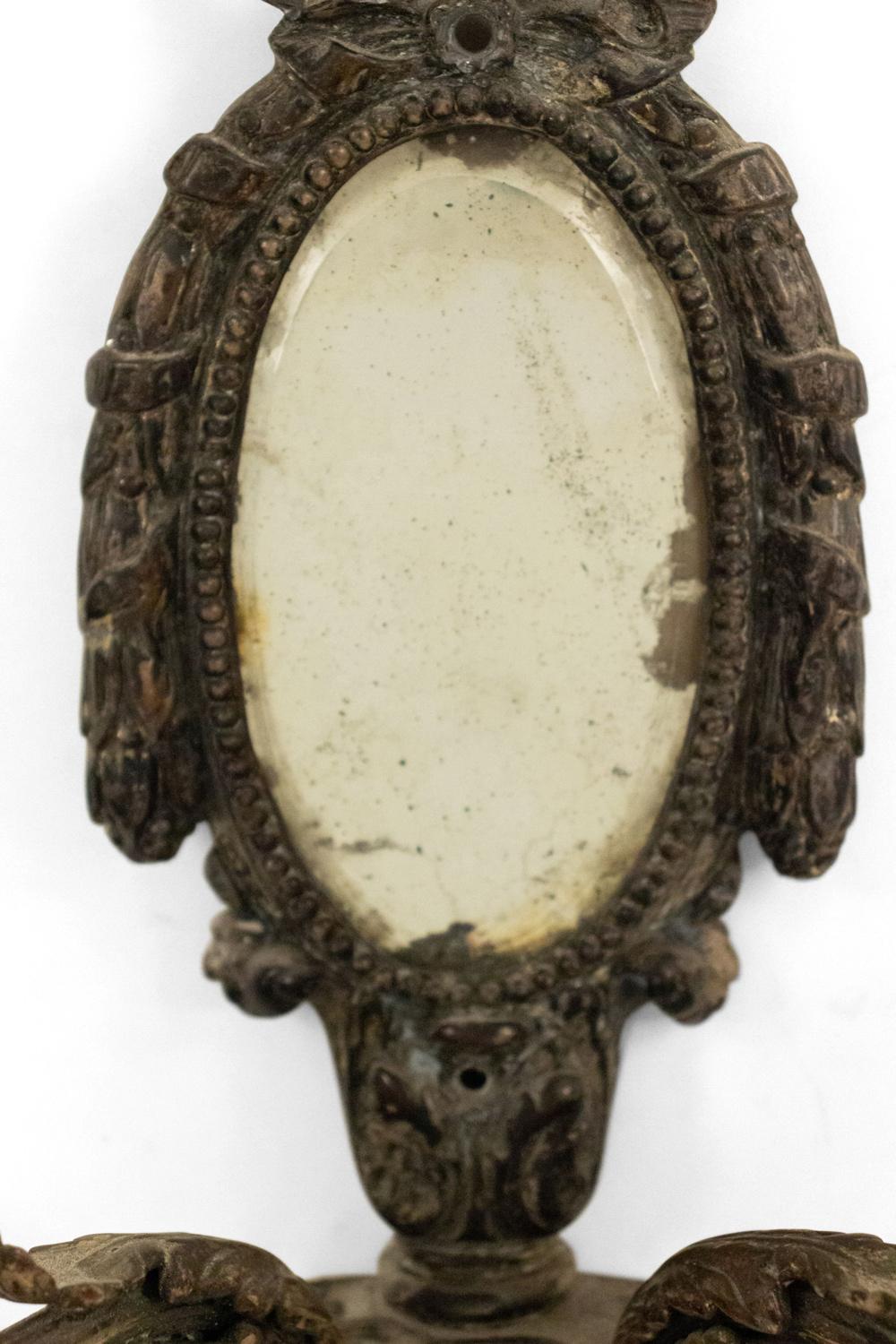 Französischer Wandleuchter aus Silberblech im Louis-XVI-Stil (19. Jahrhundert) mit zwei Schneckenarmen, ovalem Spiegel und Kranzmotiv mit Bogenknoten.