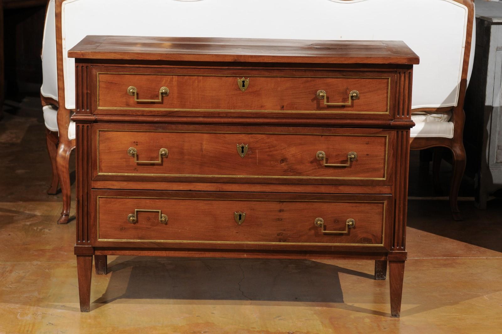 A French Louis XVI style walnut three-drawer commode from the first half of the 19th century with brass accents and fluted motifs. This French walnut commode features a rectangular top sitting above three graduated drawers. Each drawer is dovetailed