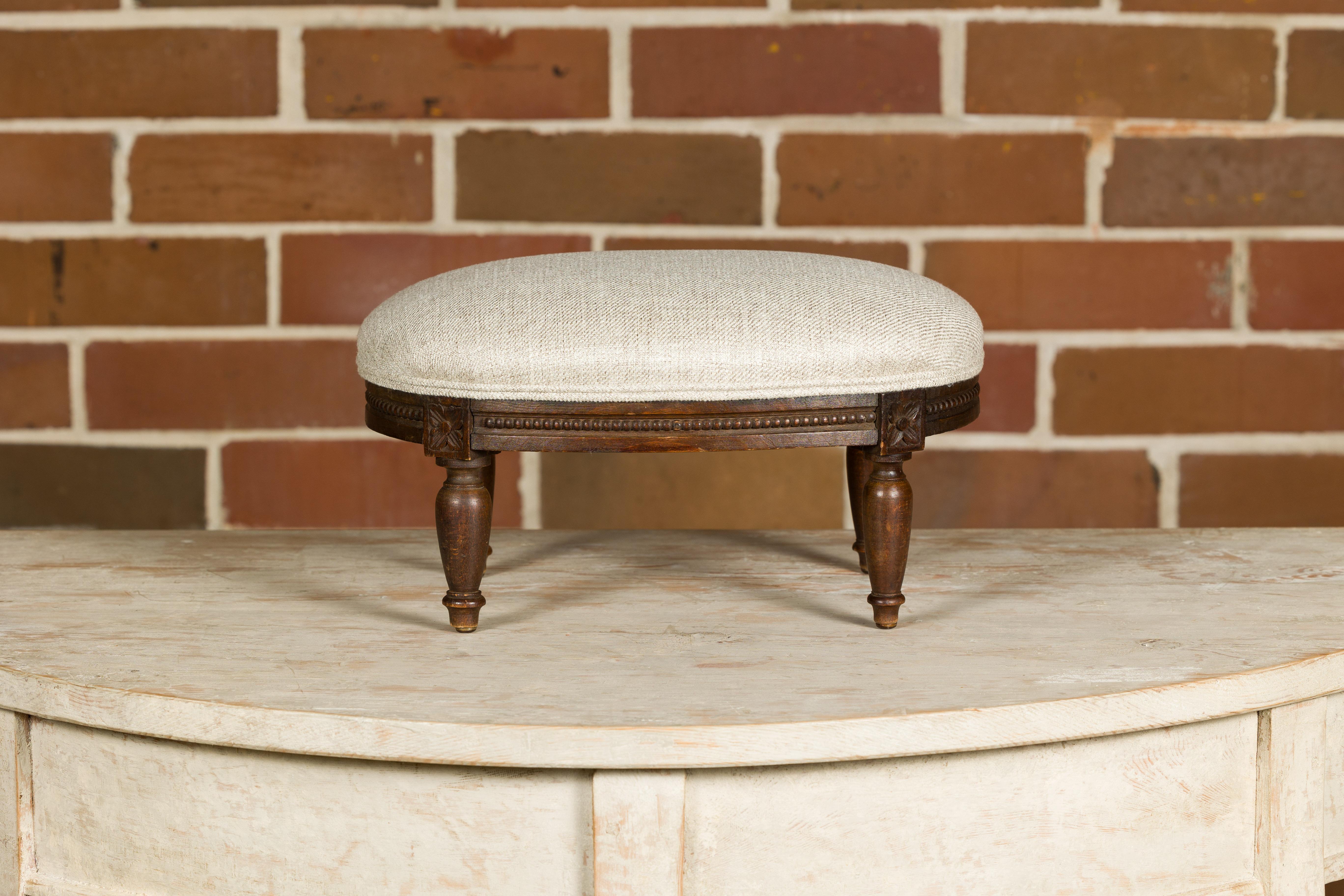 A French Louis XVI style wooden footstool from circa 1900 with carved rosettes, beads and turned legs. This French Louis XVI style wooden footstool, dating back to the early 20th century, exudes timeless elegance and craftsmanship. Its design is a