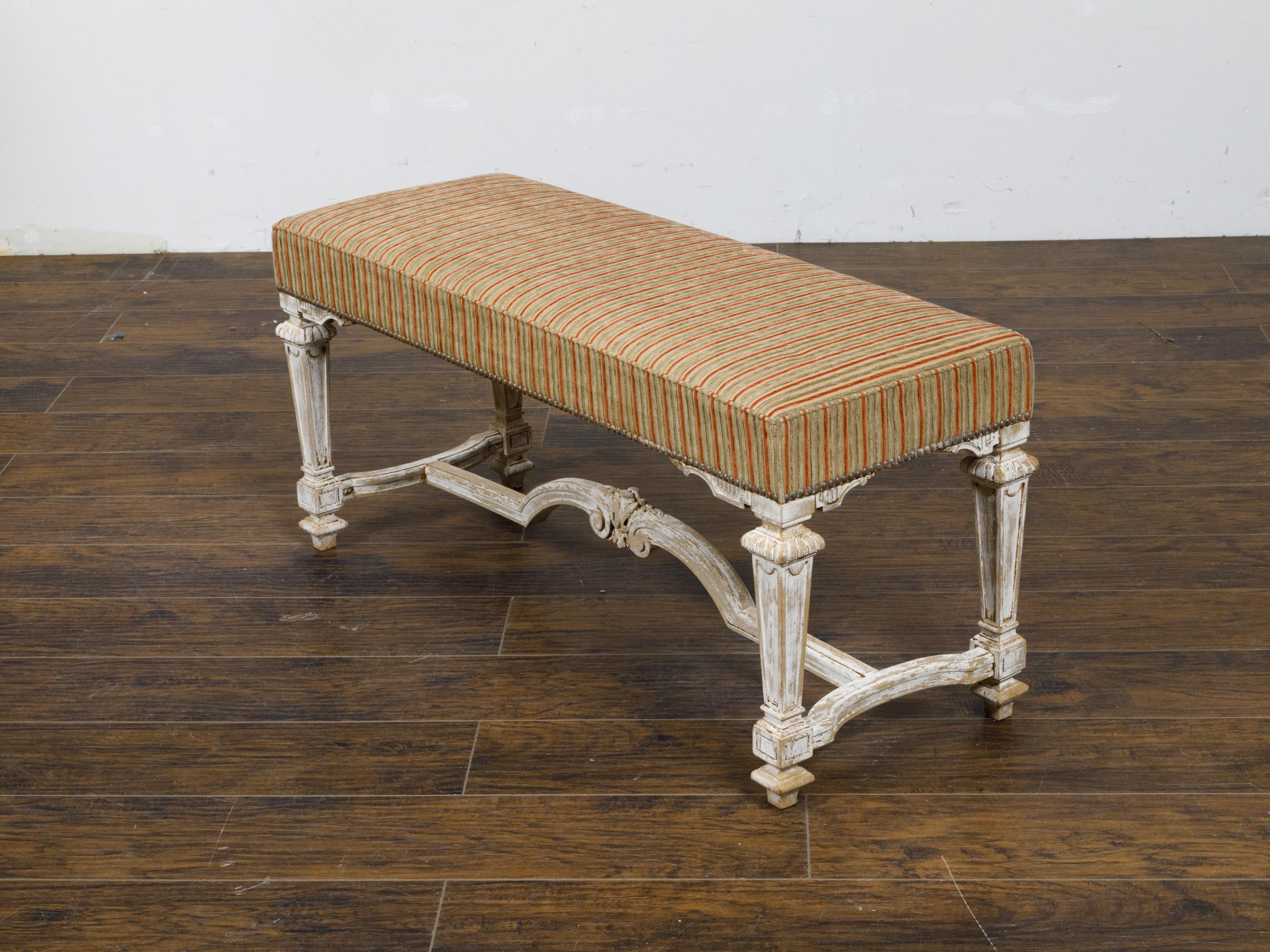 A French Louis XVI style painted wood bench from circa 1950 with off-white distressed painted finish, tapered legs and carved cross-stretcher. This exquisite French Louis XVI style painted wood bench, circa 1950, offers an air of refined elegance