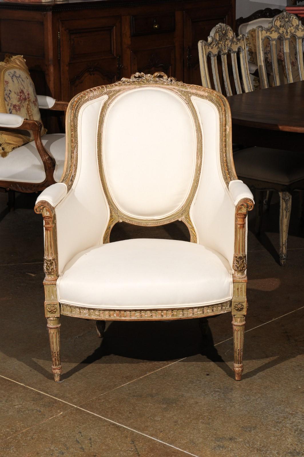 A French Louis XVI style bergère chair from the 19th century, with distressed painted finish, fluted accents, ribbon carved motif and new upholstery. Born in France during the 19th century, this exquisite armchair presents the stylistic
