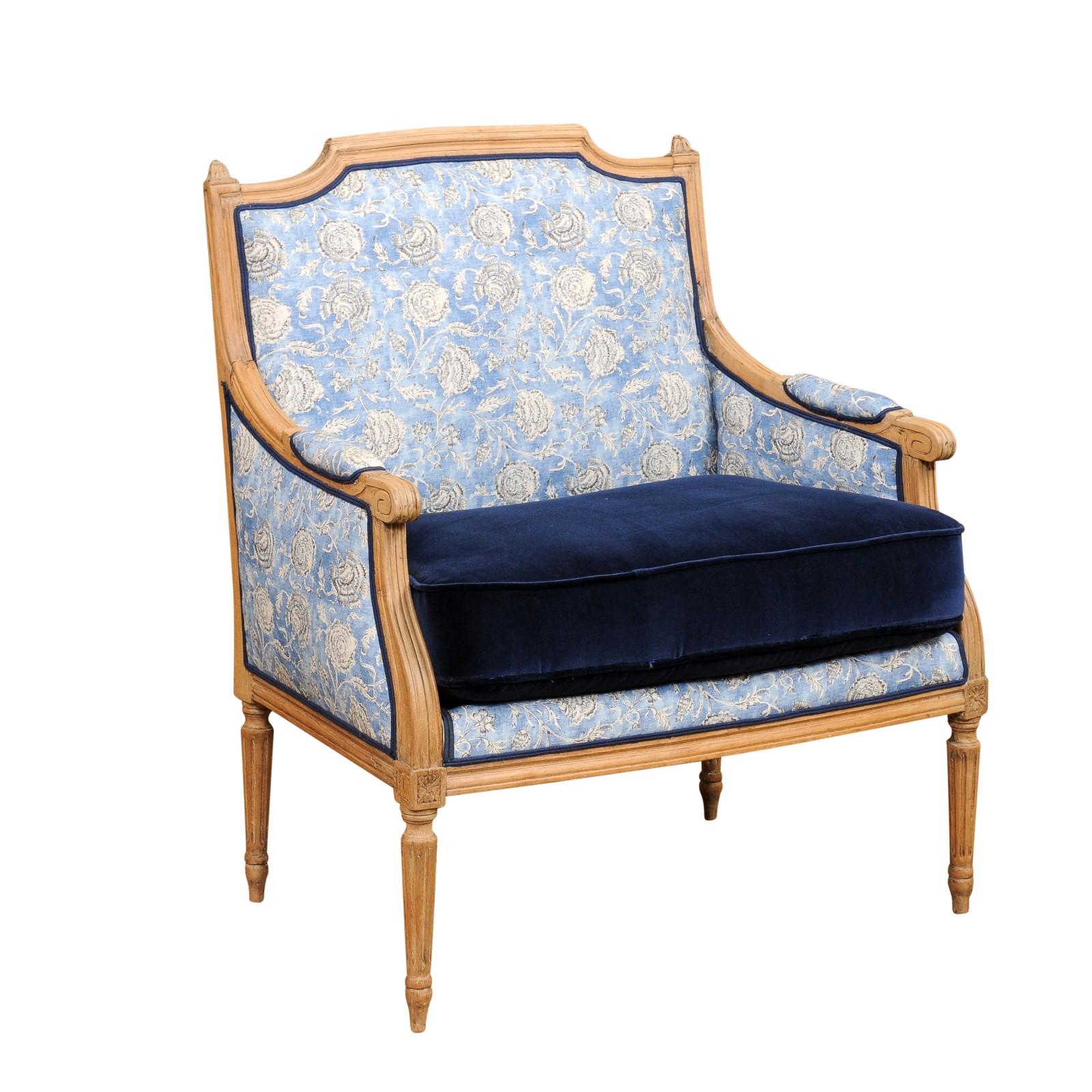 A French Louis XVI style bleached walnut marquise bergère chair from the 19th century, with slanted rectangular back, petite carved finials, scrolling knuckles on the arms, fluted legs and used blue upholstery in fair condition with custom velvet