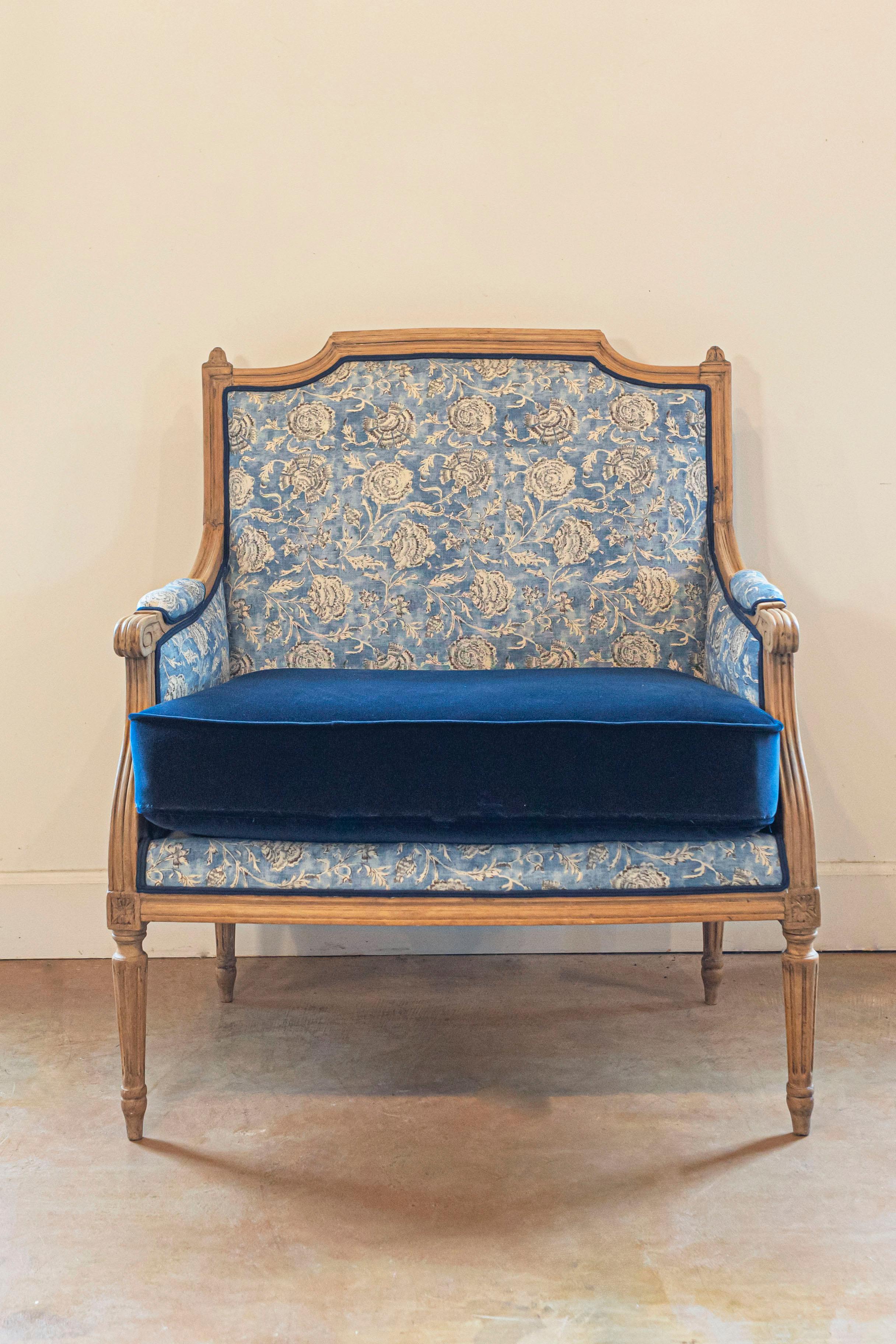 A French Louis XVI style bleached walnut marquise bergère chair from the 19th century, with slanted rectangular back, petite carved finials, scrolling knuckles on the arms, fluted legs and used blue upholstery in fair condition with custom velvet