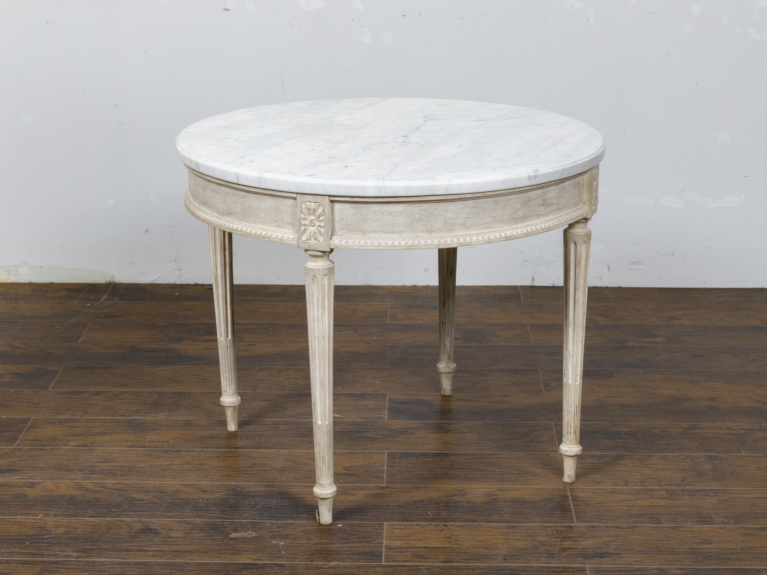 A French Louis XVI style stripped wood center table from the 19th century with circular white veined marble top, fluted legs and carved rosettes. Elevate your living space with this exquisite French Louis XVI style center table from the 19th