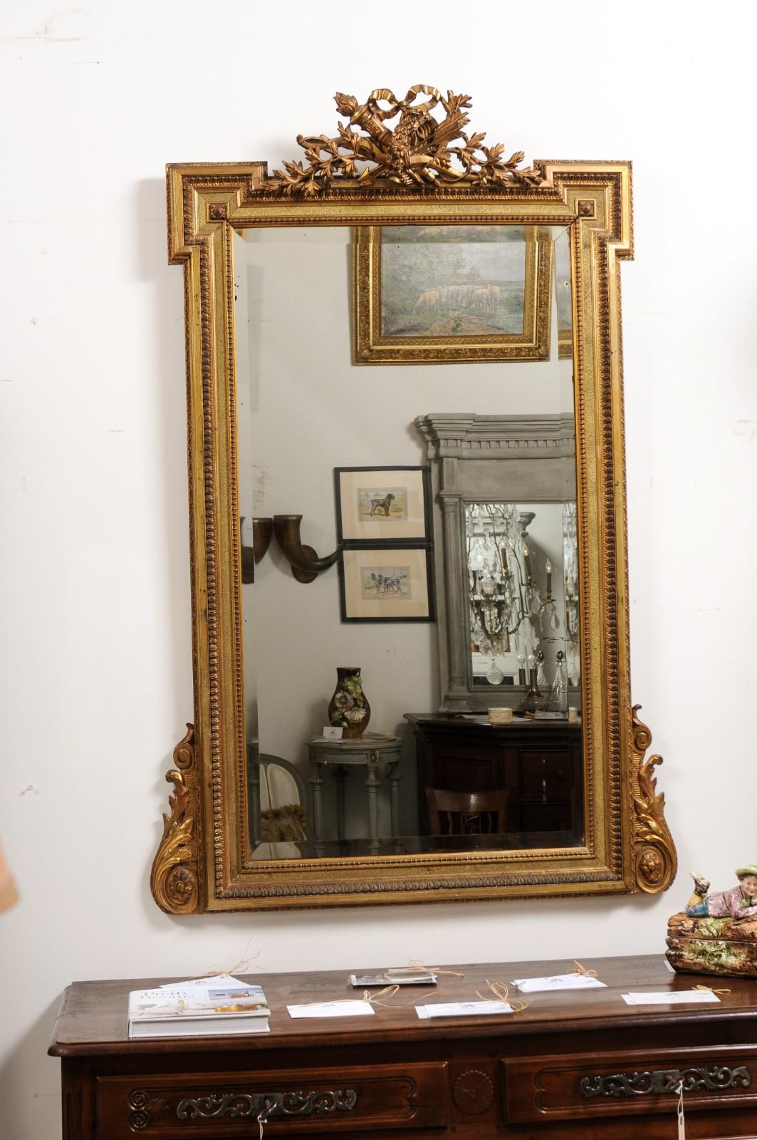 A French Louis XVI style giltwood mirror from the 19th century with hand-carved crest adorned with ribbon-tied arrows and torch motifs. Elevate your living space to the epitome of regal opulence with this resplendent French Louis XVI style giltwood