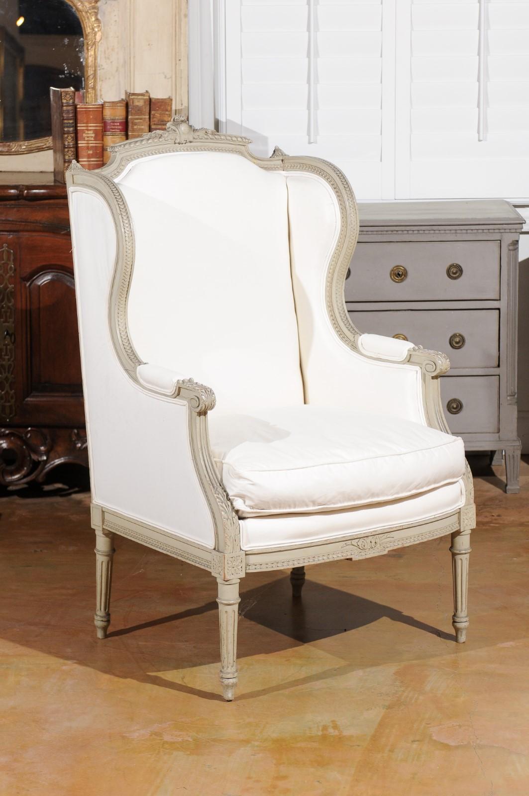 A French Louis XVI style painted wingback bergère chair from the 19th century, with carved decor and new upholstery. Born in France during the 19th century, this lovely 'bergère à oreilles' features a winged back topped with a carved cartouche in