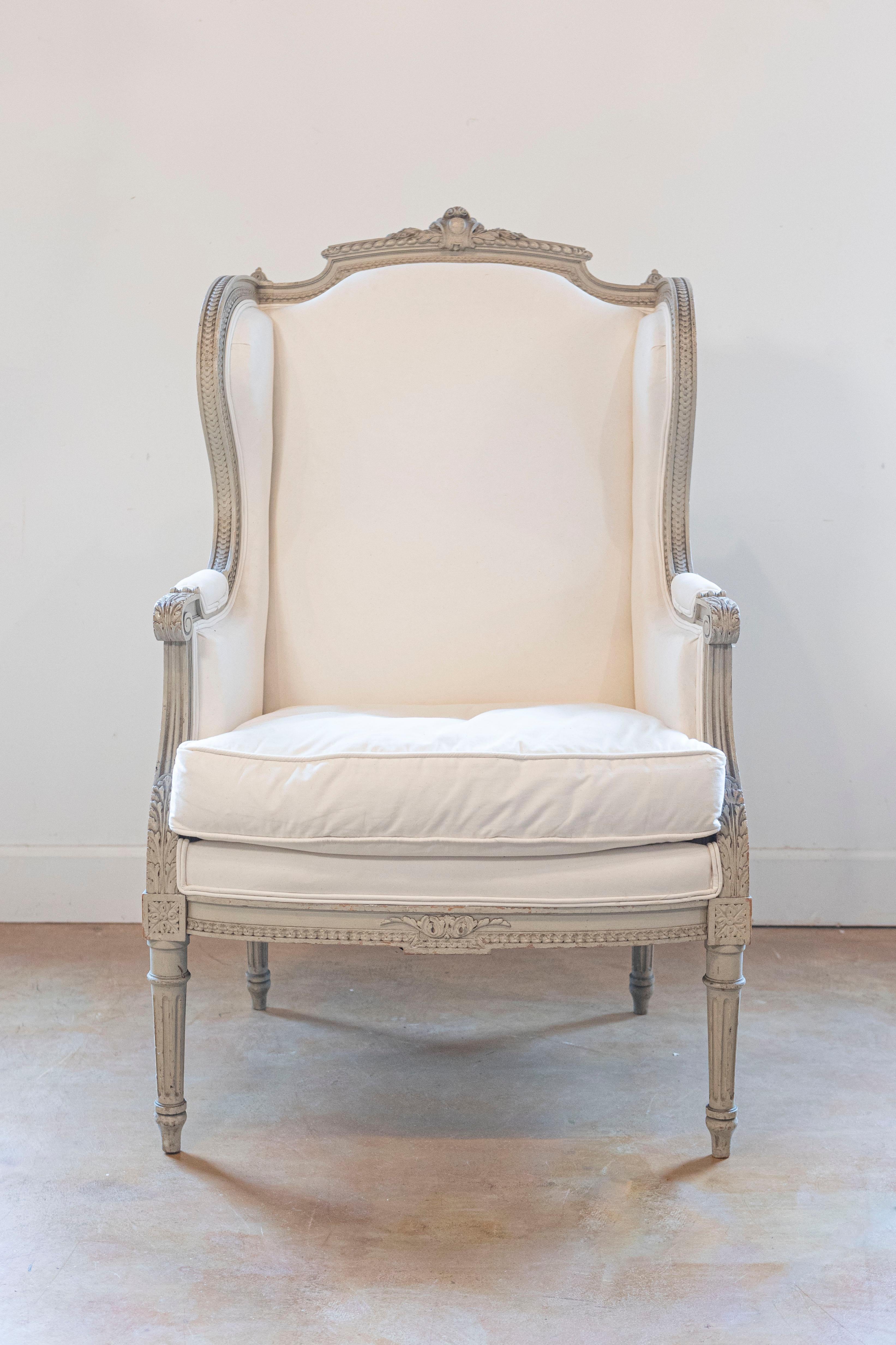 A French Louis XVI style painted wingback bergère chair from the 19th century, with carved decor and new upholstery. Born in France during the 19th century, this lovely 'bergère à oreilles' features a winged back topped with a carved cartouche in