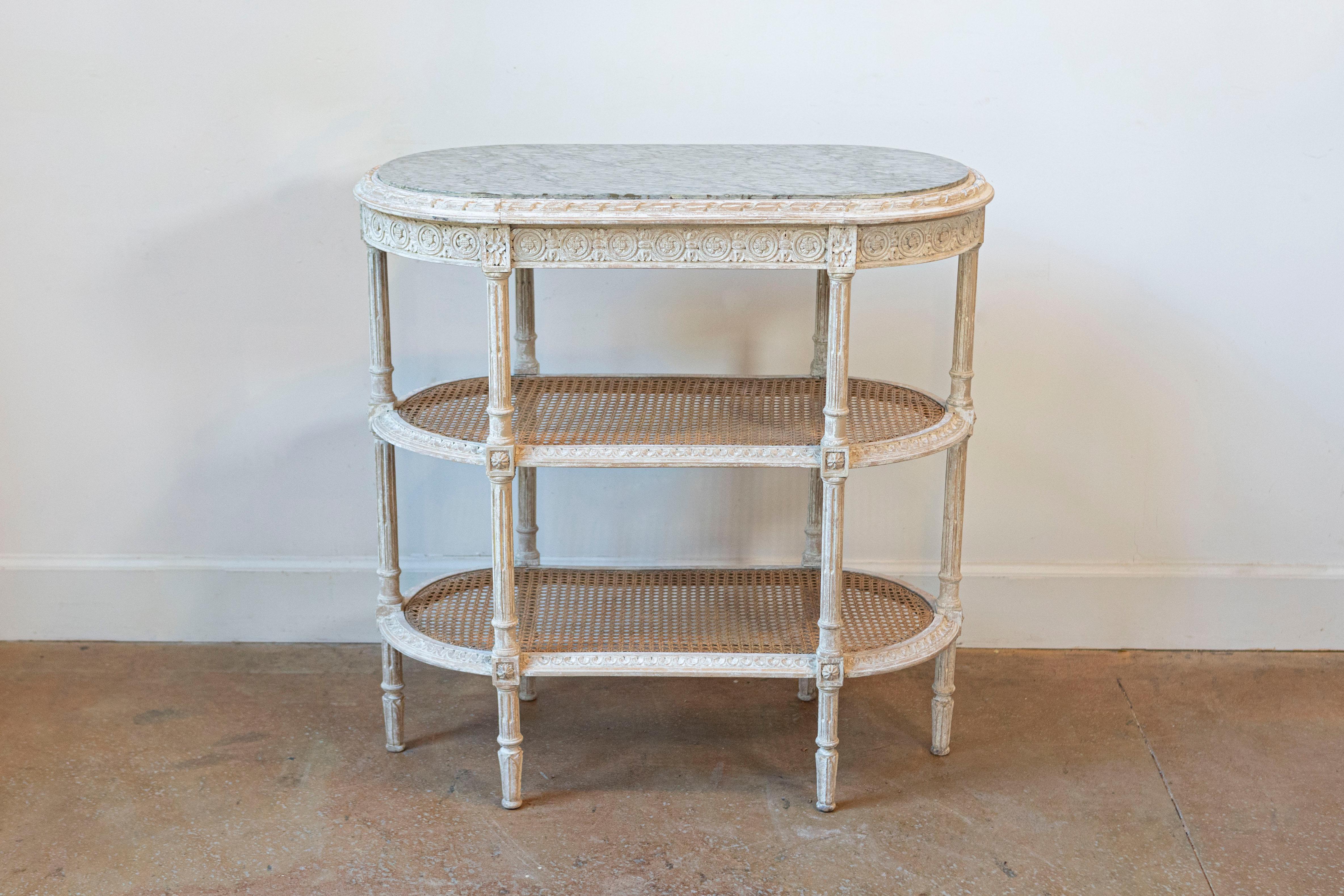 A French Louis XVI style painted wood tiered side table from the 19th century with oval marble top, carved décor showcasing medallions, rosettes and fluted legs, as well as two shelves with caning. This 19th-century French Louis XVI style tiered
