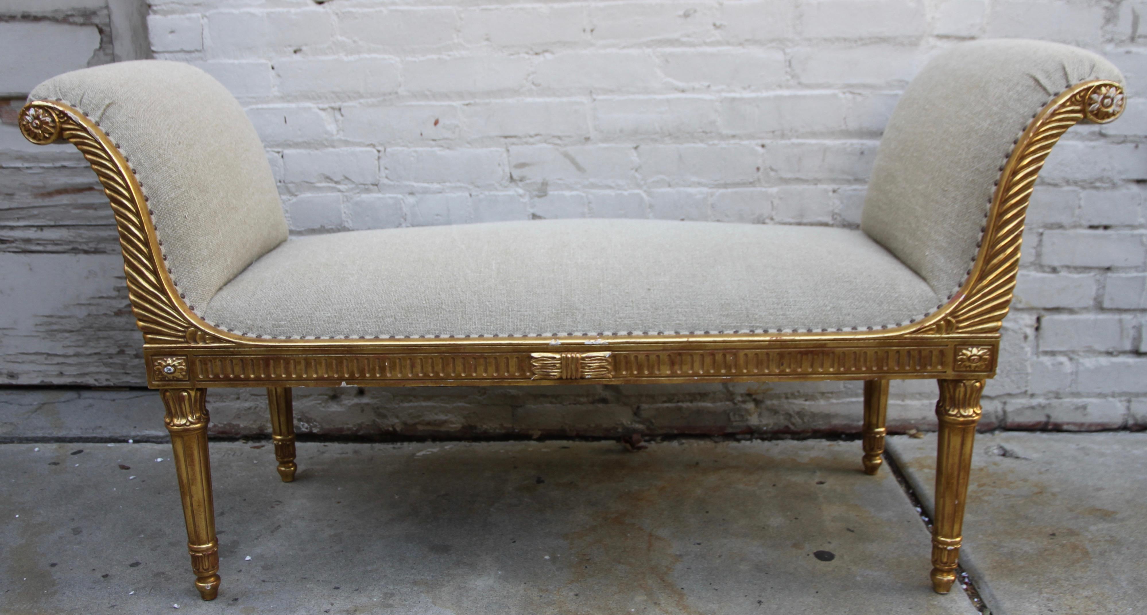 French Louis XVI neoclassical style 22-karat giltwood bench. The elegant piece stands on four fluted legs and is newly upholstered in a washed linen. Antique colored brass nailhead trim detail.