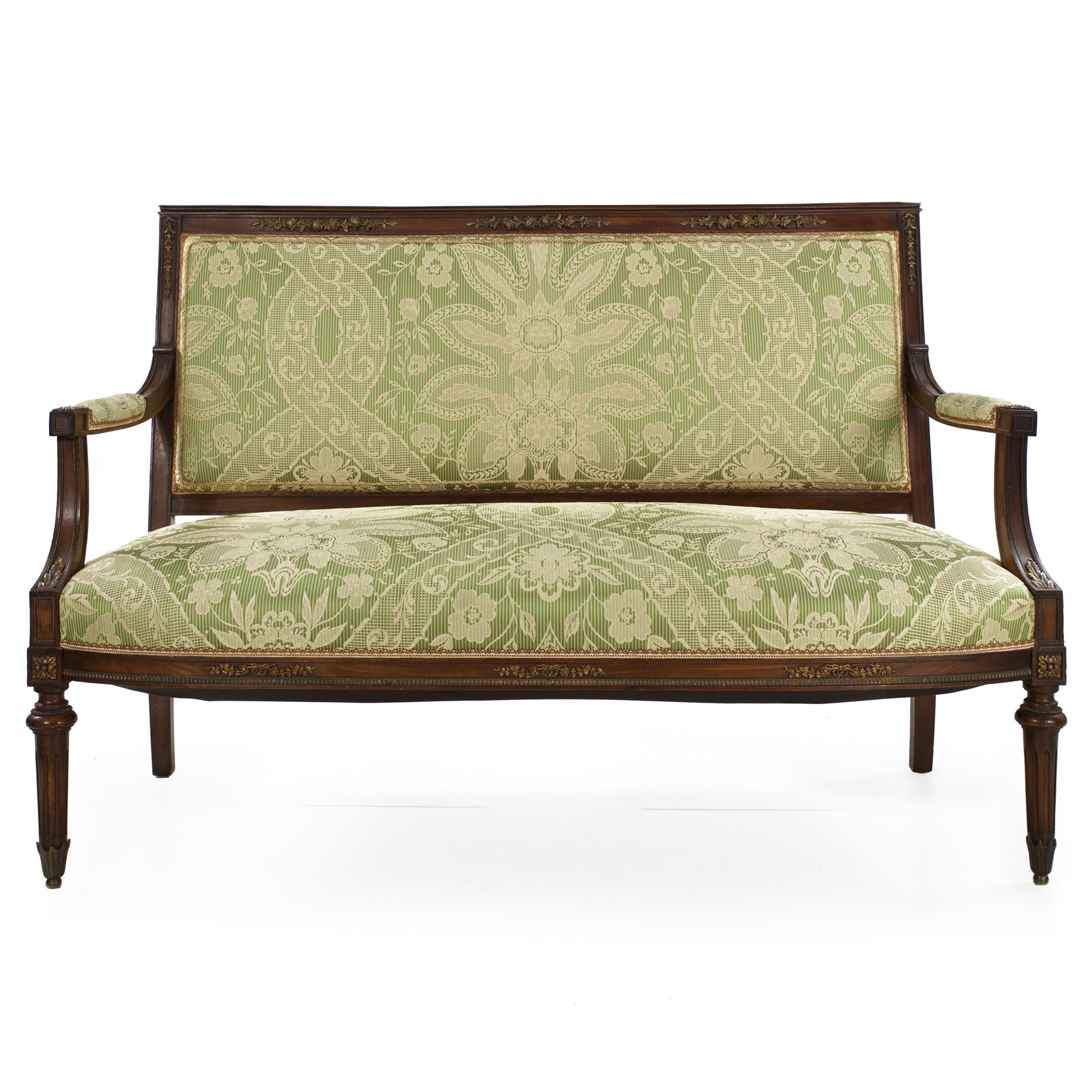 An attractive early 20th century settee in the Directoire taste, it is constructed of solid walnut with a well-developed and oxidized surface patina throughout. The crest is symmetrically embellished with vertical floral cast bronze mounts with a