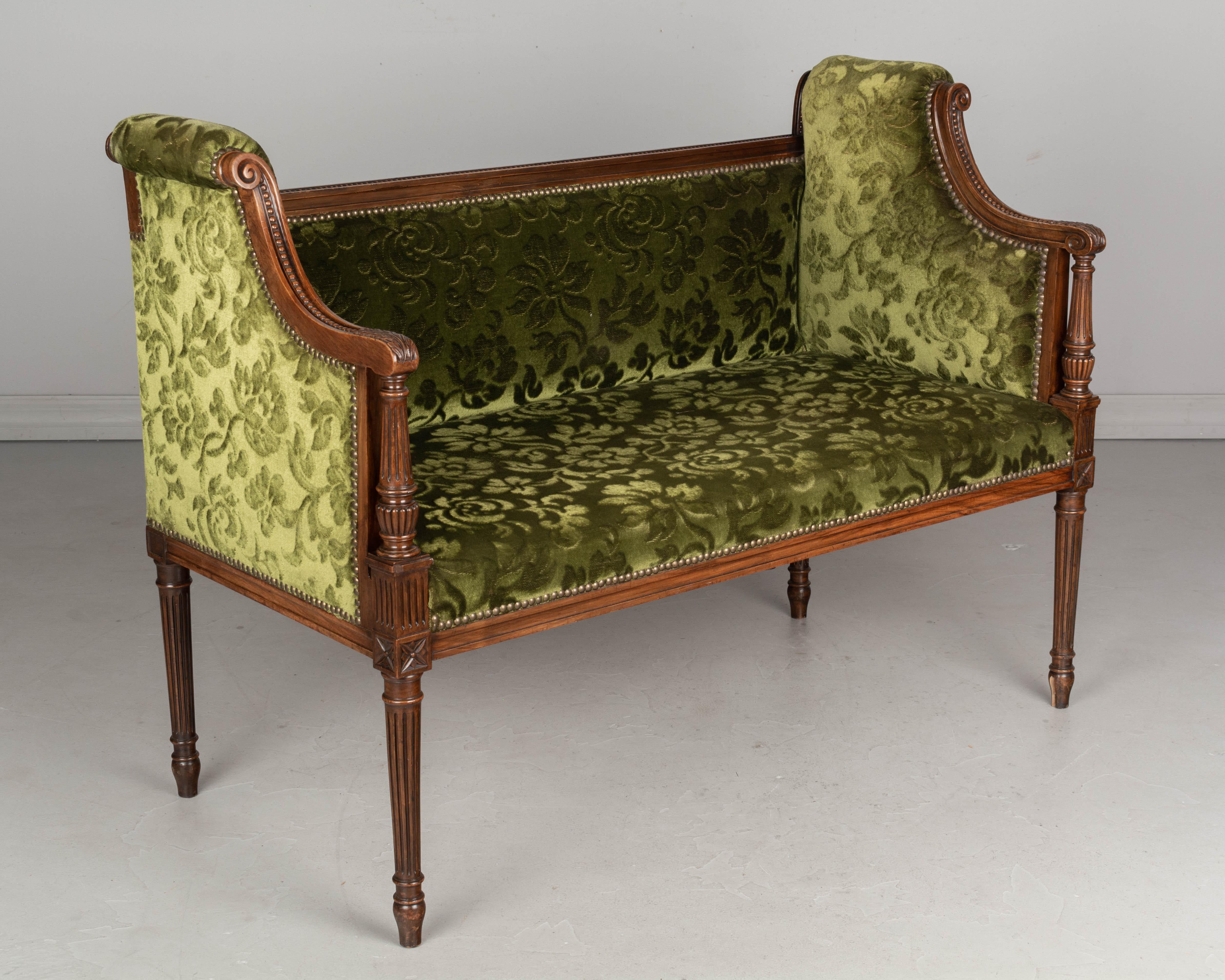 A Louis XVI style small banquette or bench with walnut frame. Good proportions and nice carved details including fluted legs and columns and sloped arms with scrolls. Old green velvet brocade upholstery is in serviceable condition. Circa 1900-1910.
