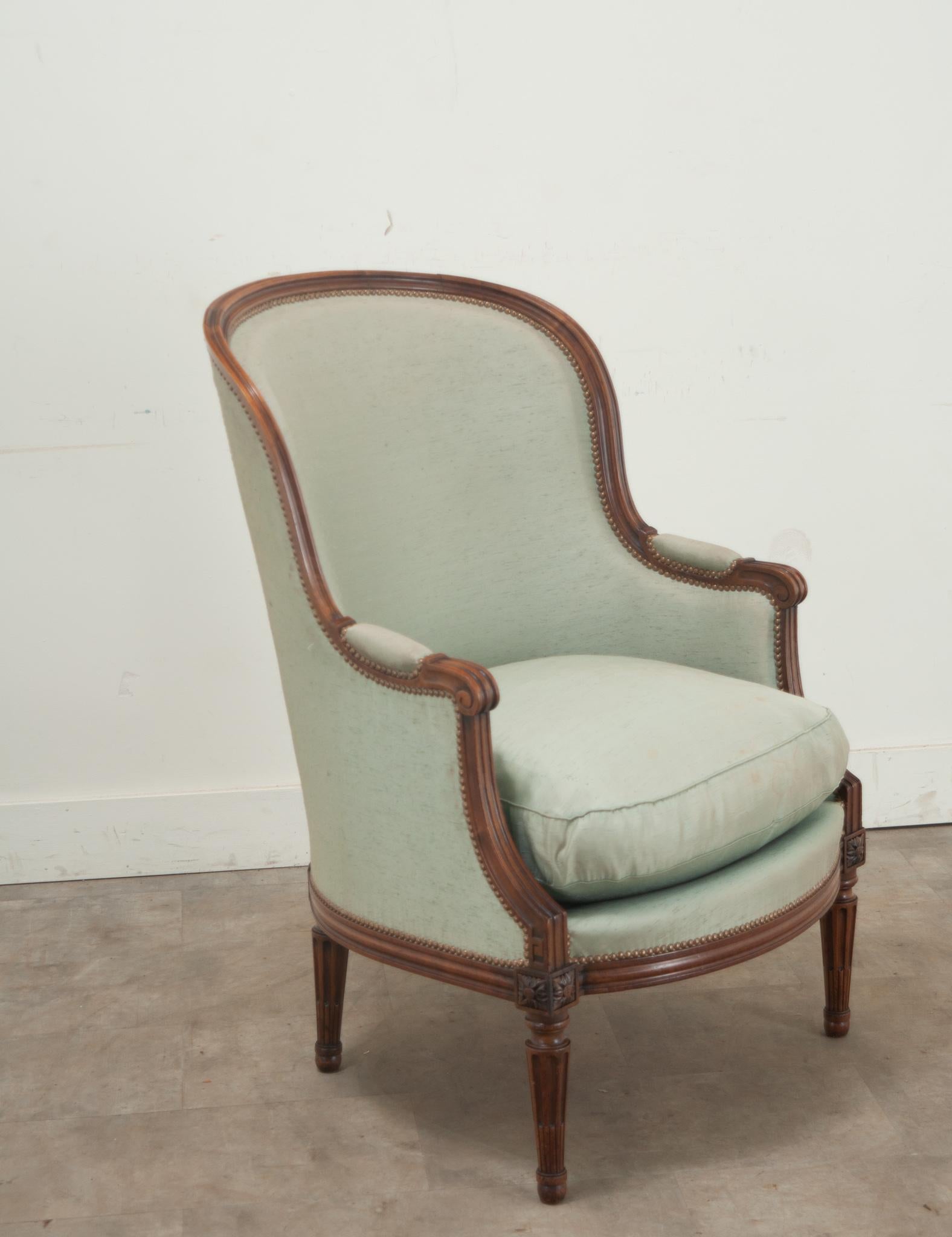 A single French Louis XVI style walnut bergere. This barrel back bergere has a walnut frame and is upholstered in a worn silk green upholstery (there is some staining on the seat cushion) with a brass nailhead trim. Louis XVI style carvings are seen