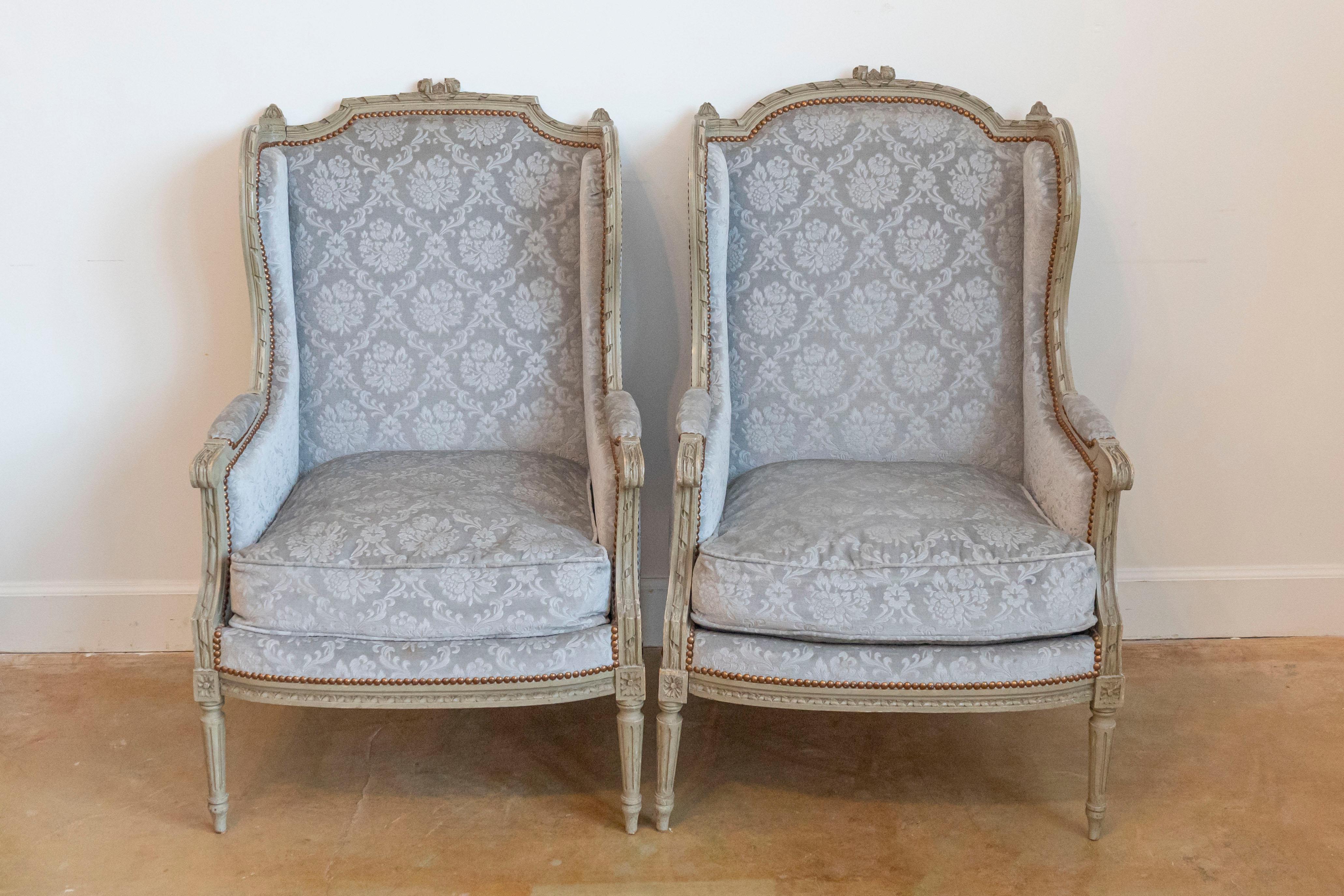 A pair of French Louis XVI style painted wood wingback bergères à oreilles from the early 20th century, with carved ribbons, twisted rails, scrolling knuckles, fluted legs and floral upholstery. Created in France during the Turn of the Century which