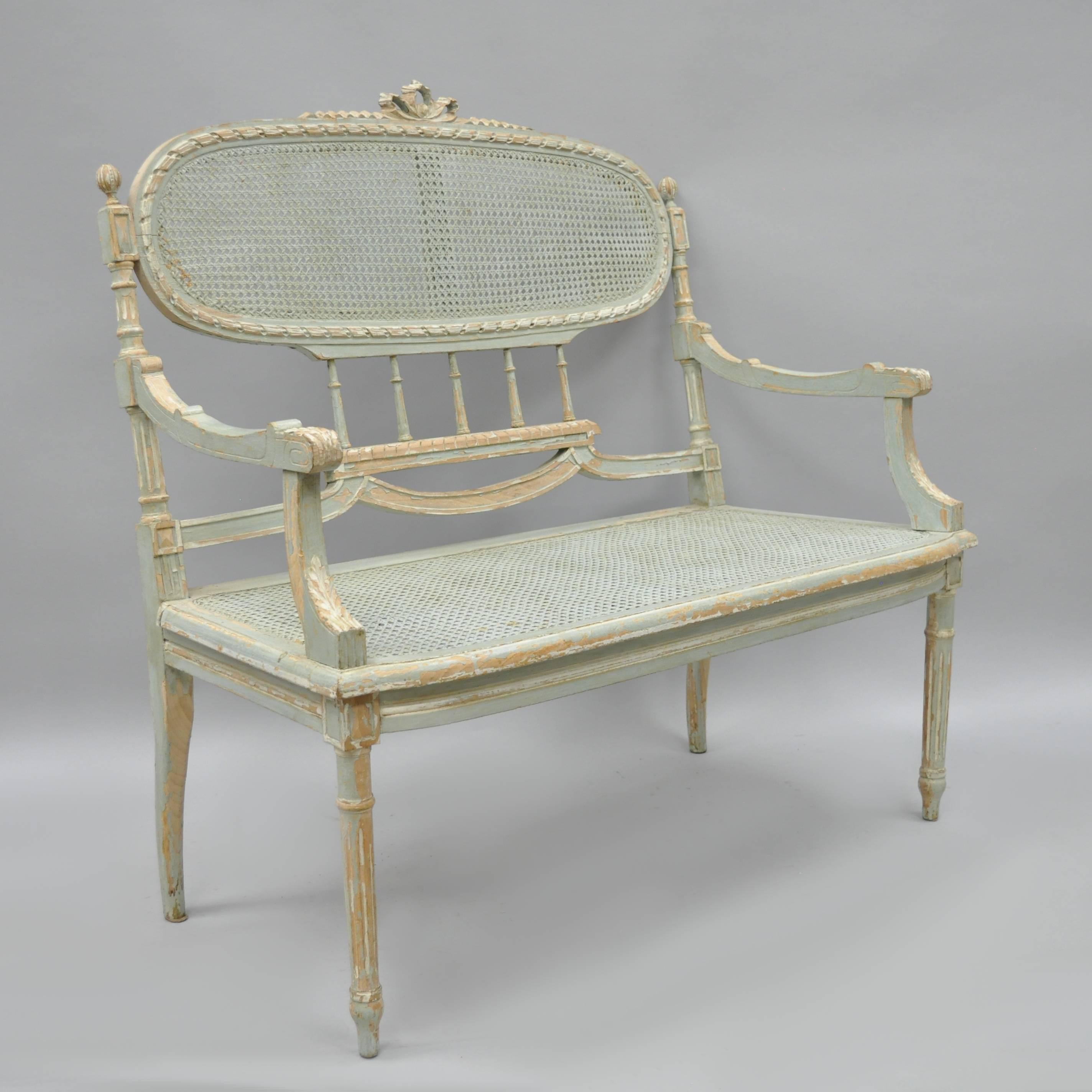 Distress painted French Louis XVI style settee. Chair is constructed of solid wood with reeded and tapered legs, carved bow tie crest and other classic French carvings throughout. Back and seat are caned and the original painted finish shows
