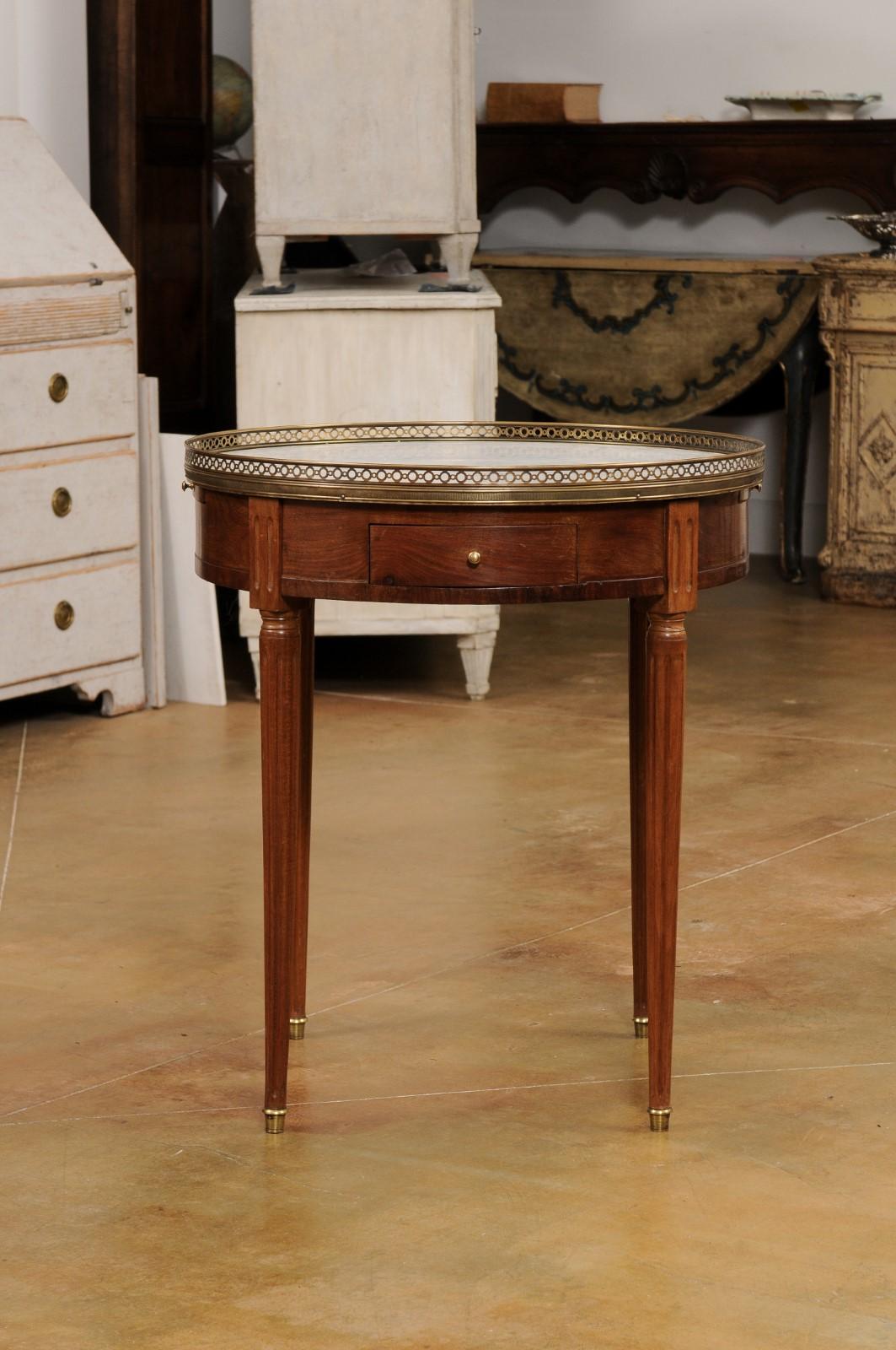 A French Louis XVI style mahogany bouillotte table from the 19th century, with white marble top, pierced brass gallery, drawers, pull-outs and fluted motifs. Created in France during the 19th century, this bouillotte table showcases the stylistic