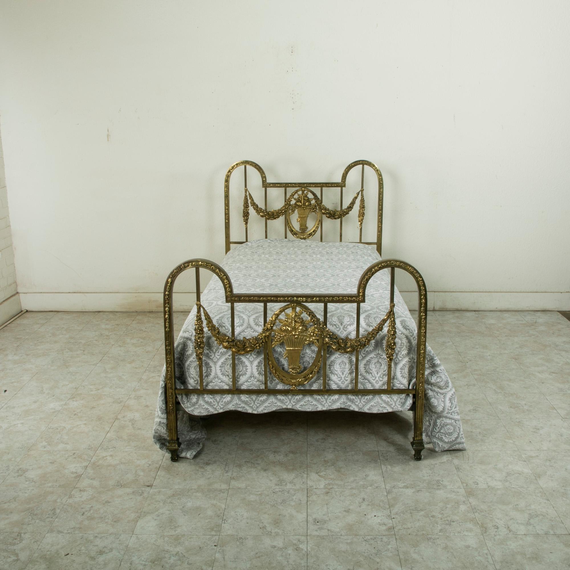 This French Louis XVI style brass bed from the turn of the twentieth century features a headboard and footboard detailed with a central basket motif, from the region of Normandy. Garlands of flowers flank the basket and join the fluted flame. The