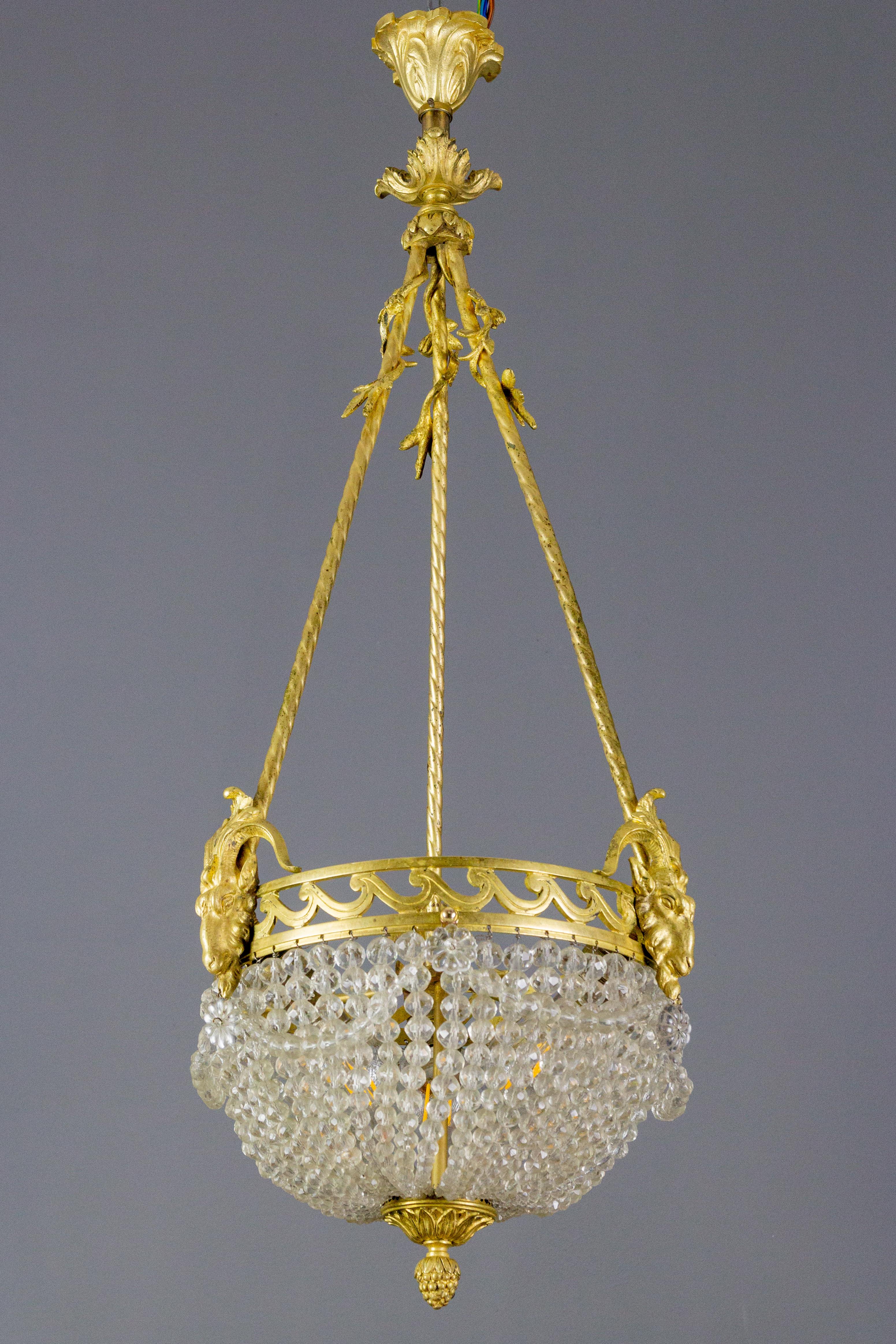 An adorable French Louis XVI style chandelier from the 1920th. The central lighting cluster is surrounded by beautiful beaded crystal garlands and an elegant rim with three ram's heads and suspended by three bronze ropes, which are decorated with