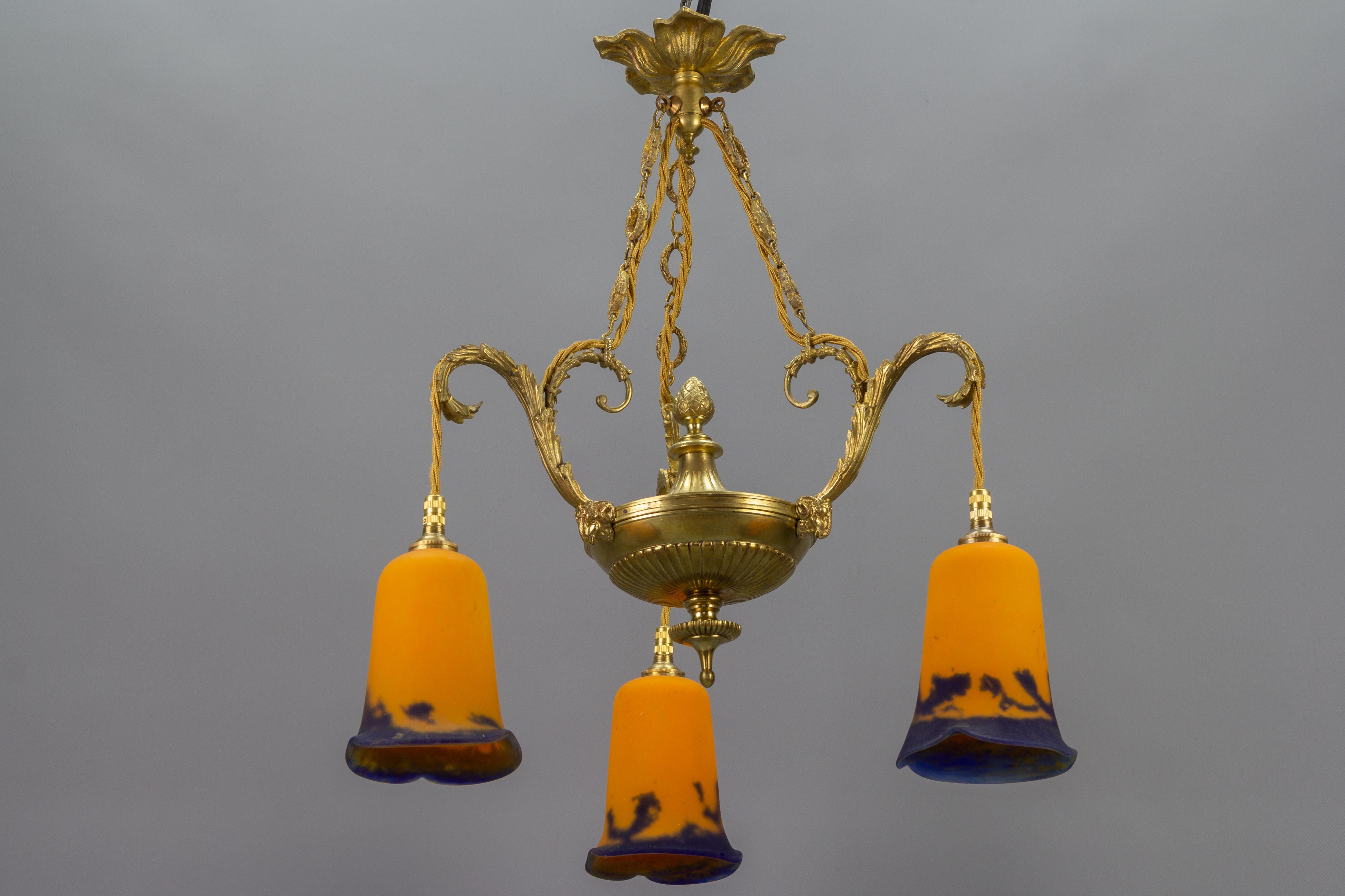 Beautiful French Louis XVI style three-light bronze chandelier with orange and blue pate de Verre glass lampshades signed ”Noverdy France” by Jean Noverdy. An ornate bronze fixture decorated with ram heads and acanthus leaf scrolls.
Three sockets