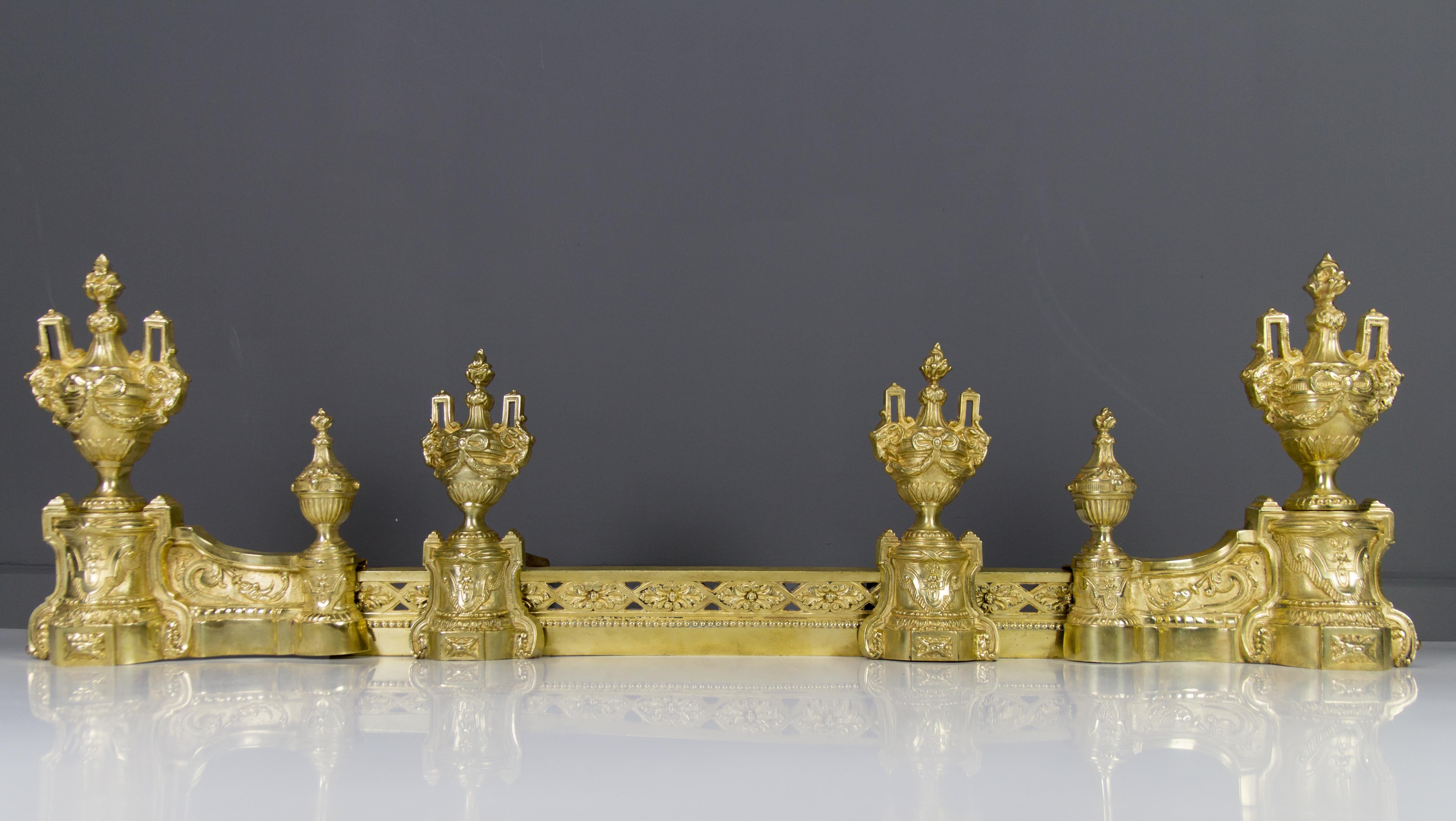 These impressive French Louis XVI style bronze andirons with their original fender date to the second half of the 1800s and are made by the famous French Bronzier Charles Casier. Richly ornate with ram heads, bows, drapes, foliate accents, urn, and