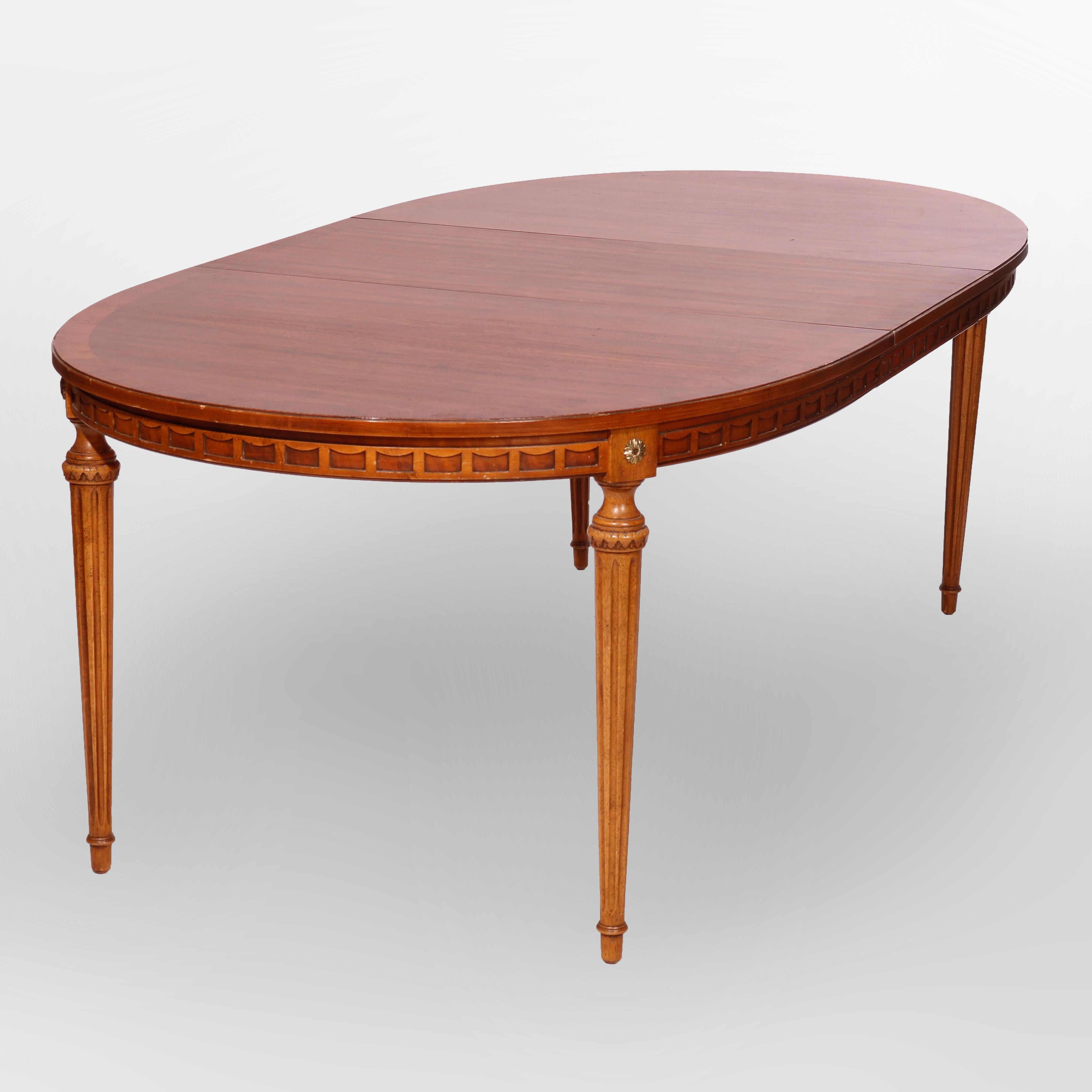 A French Louis XVI style extension dining table with two leaves offers walnut construction with cross banded oval top having carved skirt with stylized drape pattern and rosettes, raised on tapered and reeded legs, extends to accepts two leaves,