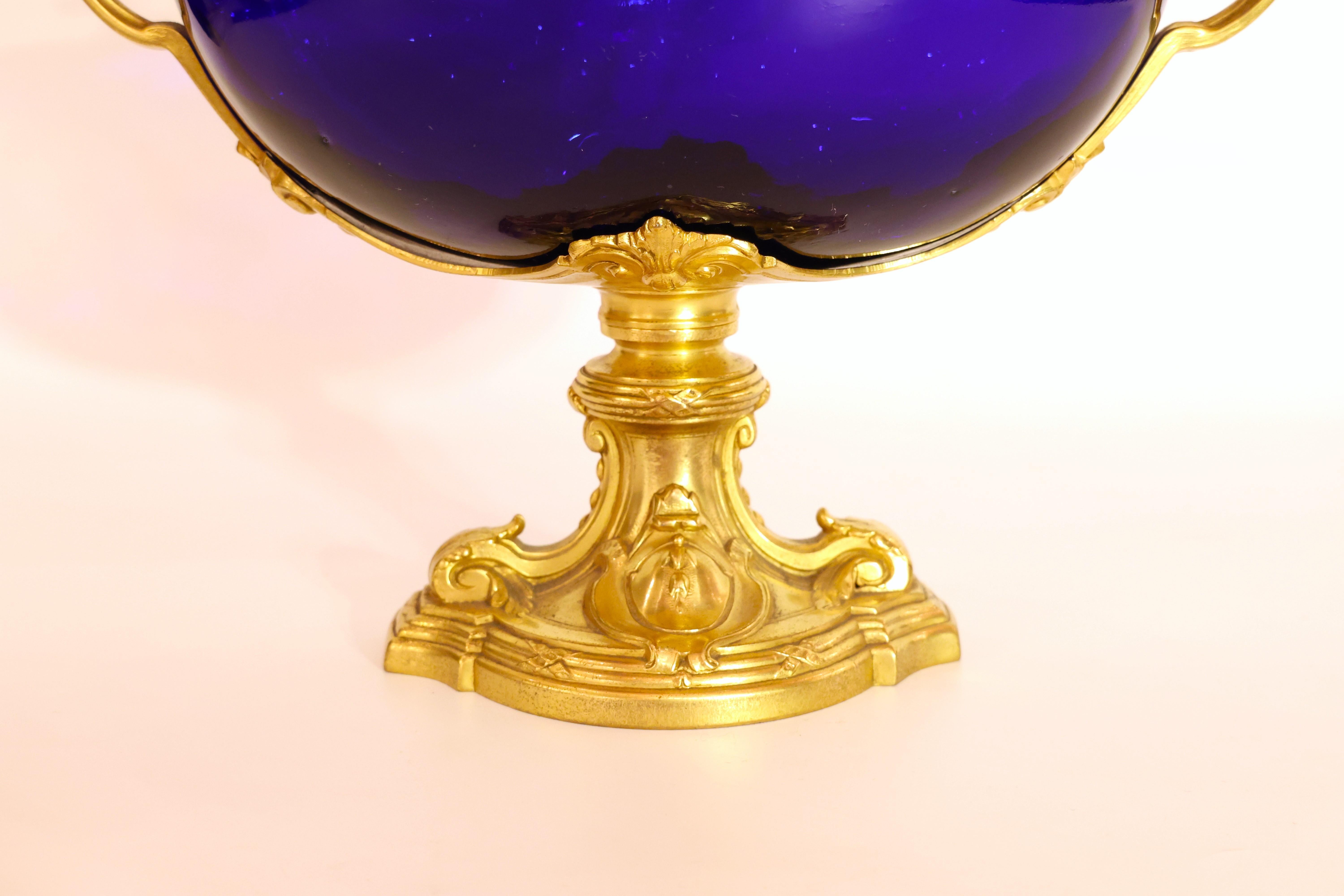 Cet exquis centre de table en bronze doré et verre bleu cobalt de style Louis XVI est surmonté d'un piédestal en bronze doré orné de rinceaux feuillagés finement ciselés, d'un cartouche central et d'accents cannelés. La base est surmontée d'une tige