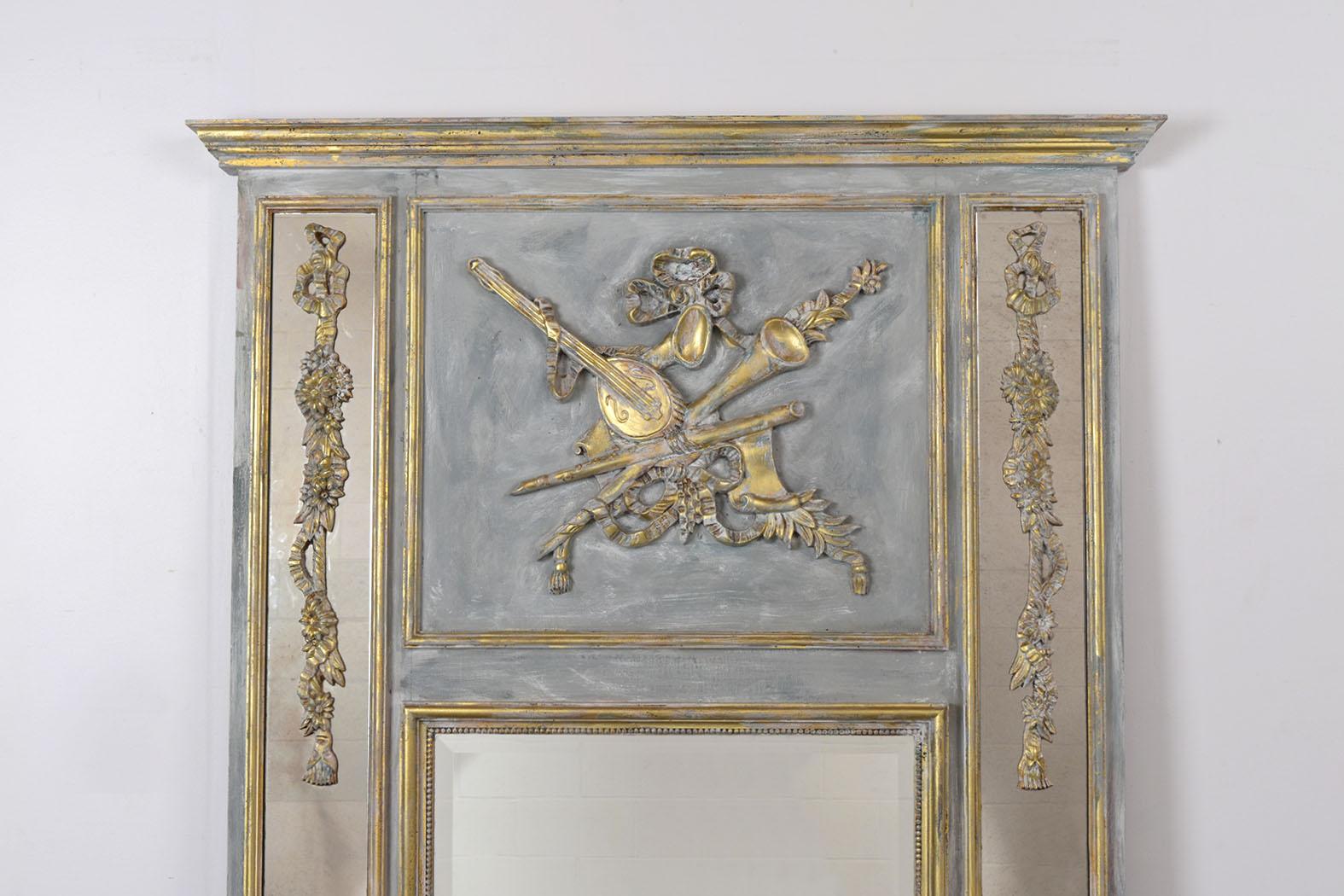French 1930s Louis XVI Style Trumeau Mirror is in good condition and features large intricately carved wood moldings of instruments, garlands flowers, and urns vases. This trumeau also has a gilt and distressed painted finish and a beveled center