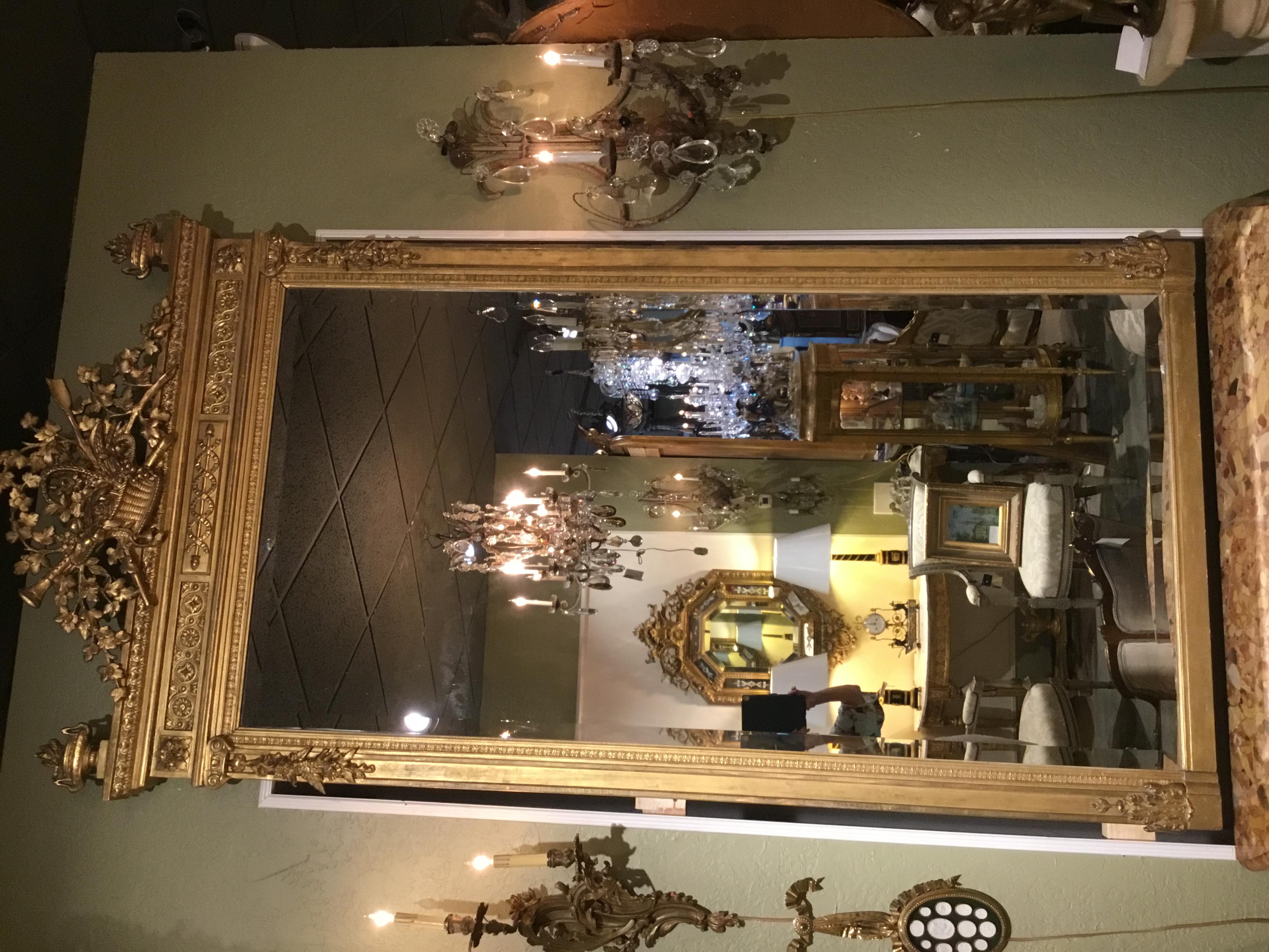 Tall and elegant giltwood 19th century Framed mirror with beveled mirror plate.
A basket with foliate and floral design with a trumpet and arrow are carved
At the center crest of this piece. Urns with a torch positioned on each of
The upper