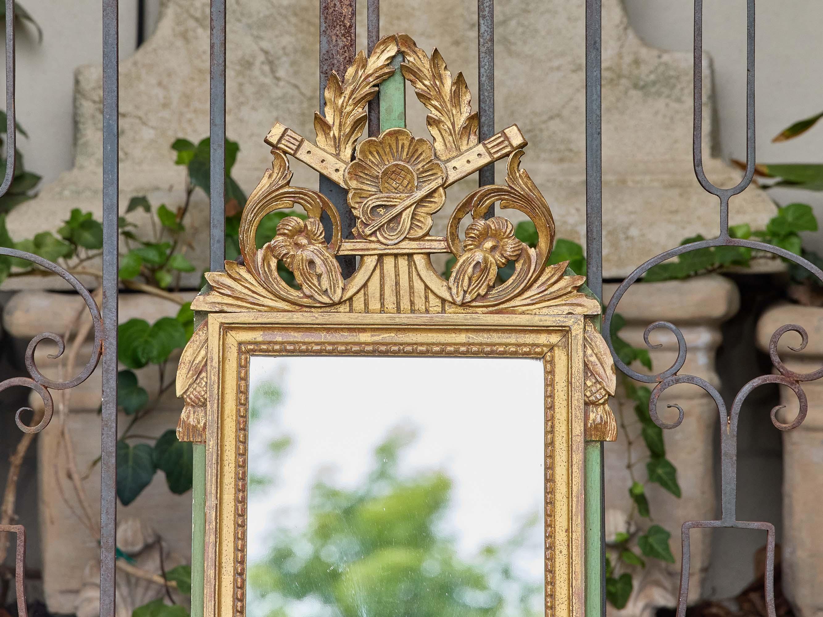Encantador espejo francés de madera dorada de estilo Luis XVI, de hacia 1920-1930, con una cresta neoclásica finamente tallada con flautas cruzadas, roleos florales y ramas de laurel. La roseta central ancla el motivo superior simétrico, mientras
