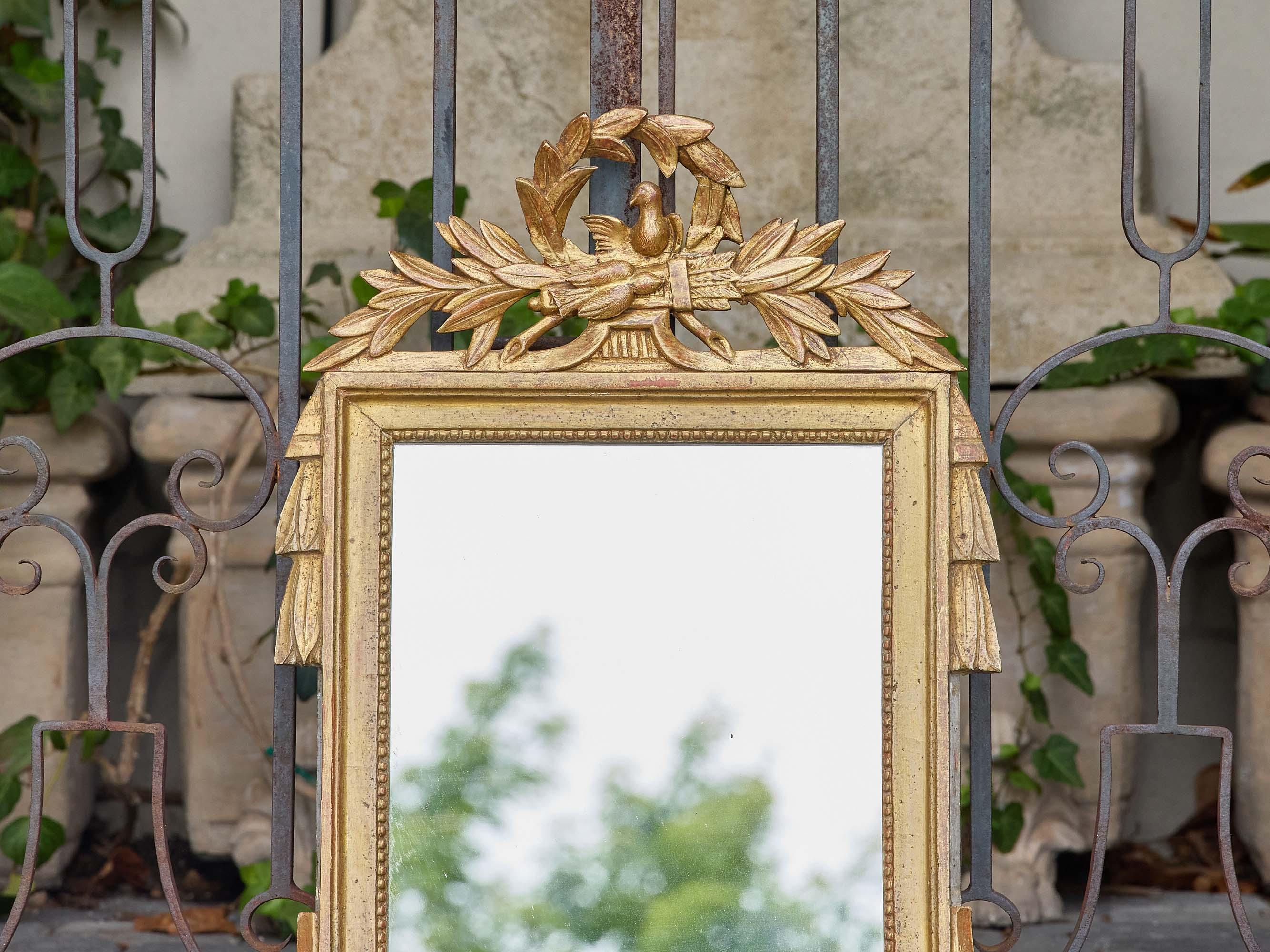This French giltwood mirror, dating to circa 1920, is crafted in the Louis XVI style and adorned with neoclassical motifs that speak to timeless elegance. The crest features a finely carved dove resting within a laurel wreath—traditional symbols of