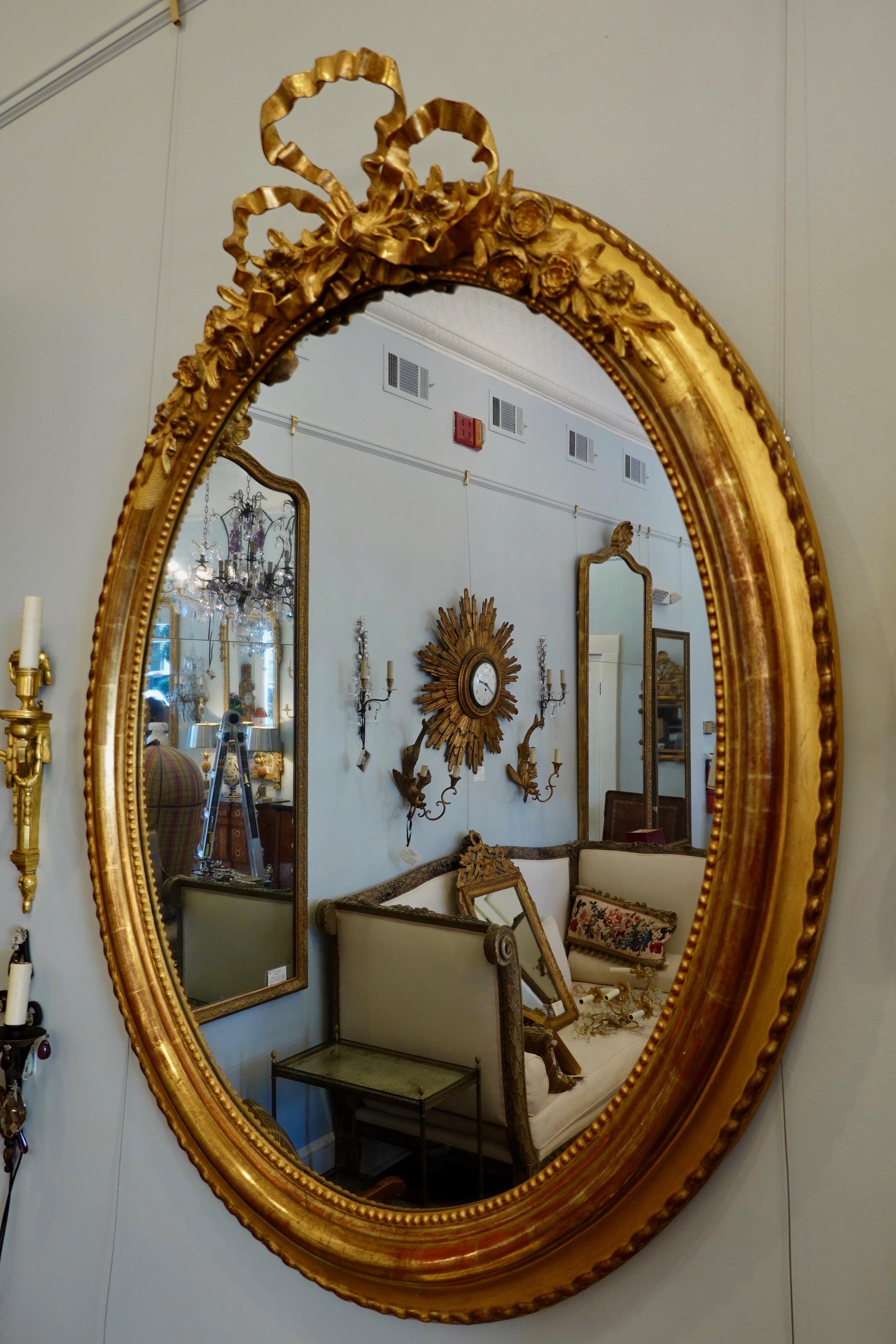 French Louis XVI style giltwood oval mirror with a large, finely-carved bow cartouche flanked by roses (circa 1830). The deeply molded frame features pearl beading on the interior perimeter and twisted molding on the exterior. Mercury glass and