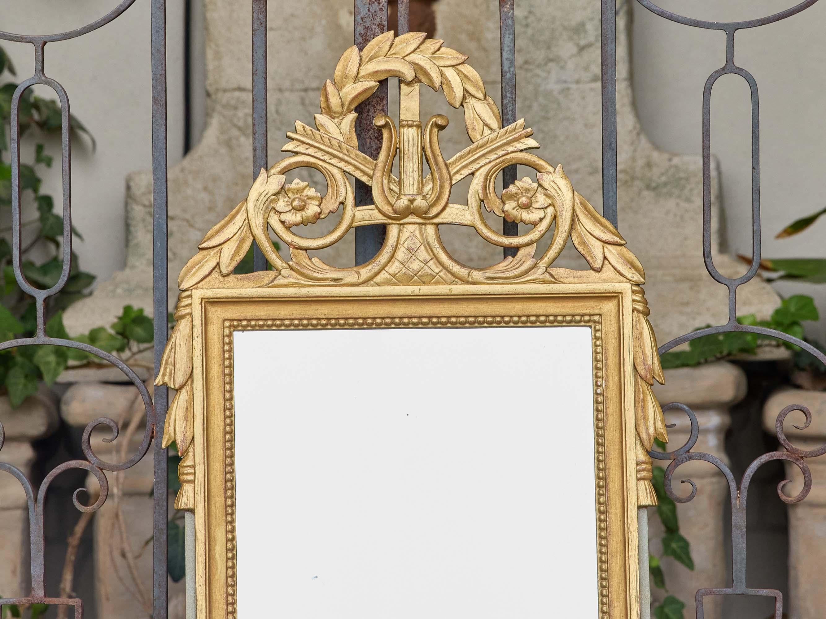This French giltwood mirror, dating to circa 1940, showcases the refined symmetry and ornamentation typical of the Louis XVI style. Crowned with a delicately carved lyre surrounded by laurel leaves and scrolls, the crest evokes classical themes of