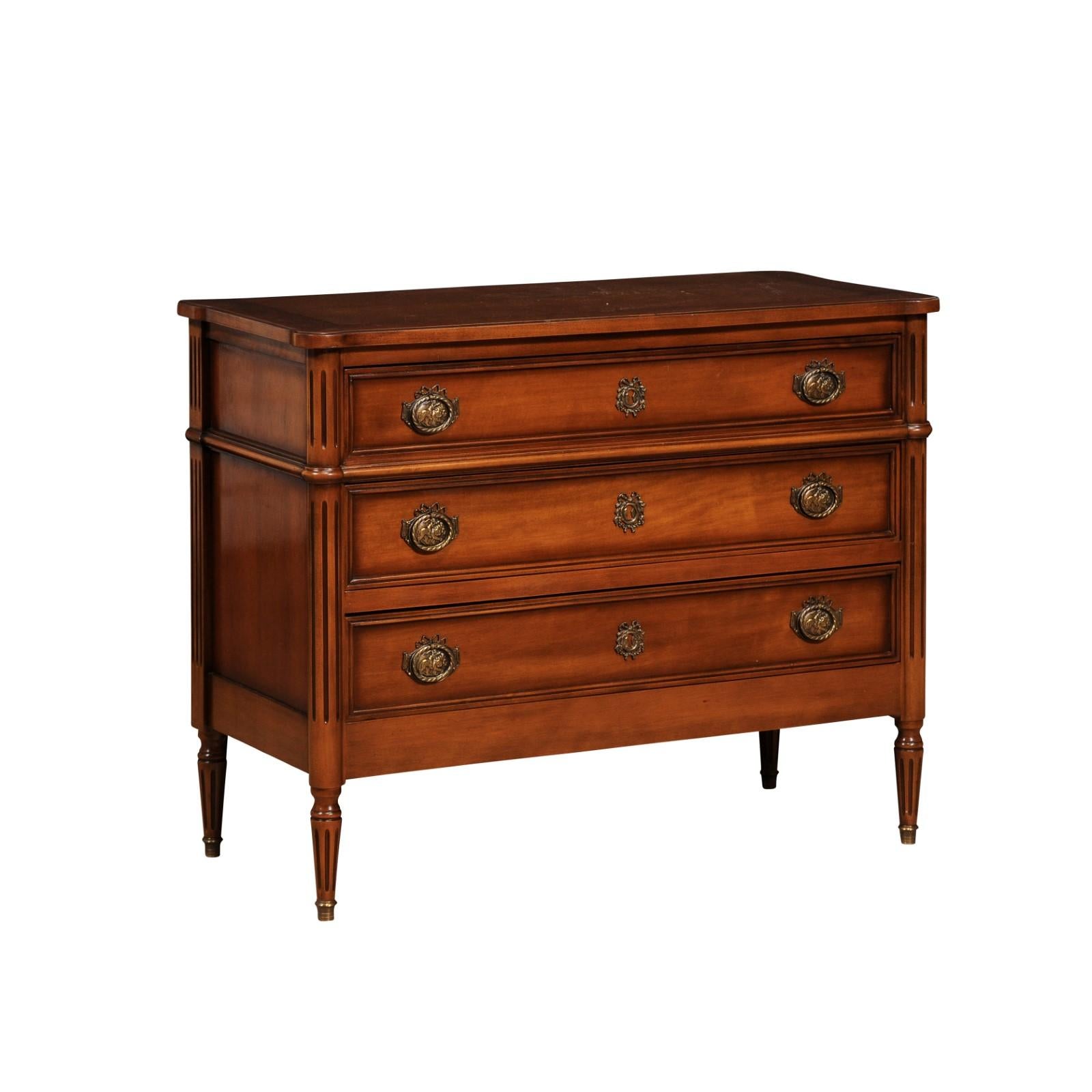 A French Louis XVI style light walnut commode from the 20th century with three drawers, fluted side posts and legs, ornate hardware. This French Louis XVI style light walnut commode from the 20th century elegantly captures the refined aesthetics of