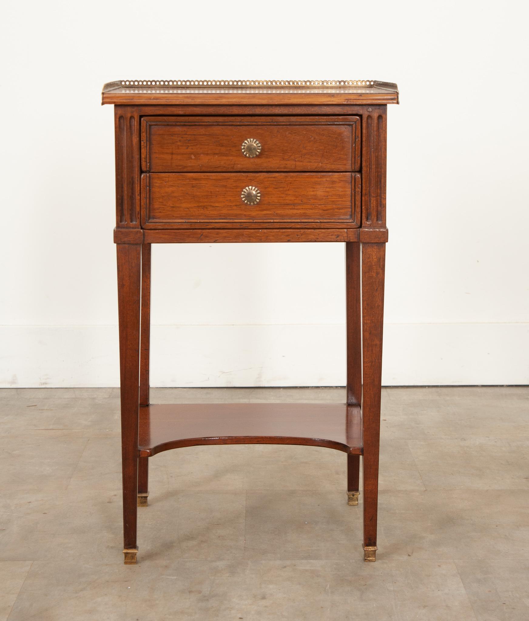 This charming mahogany bedside table was crafted in 19th century France circa 1870. A breakfront pierced brass gallery surrounds the well patinated smooth wood top. Two trimmed drawers flanked by fluted corners are fixed with small starburst brass