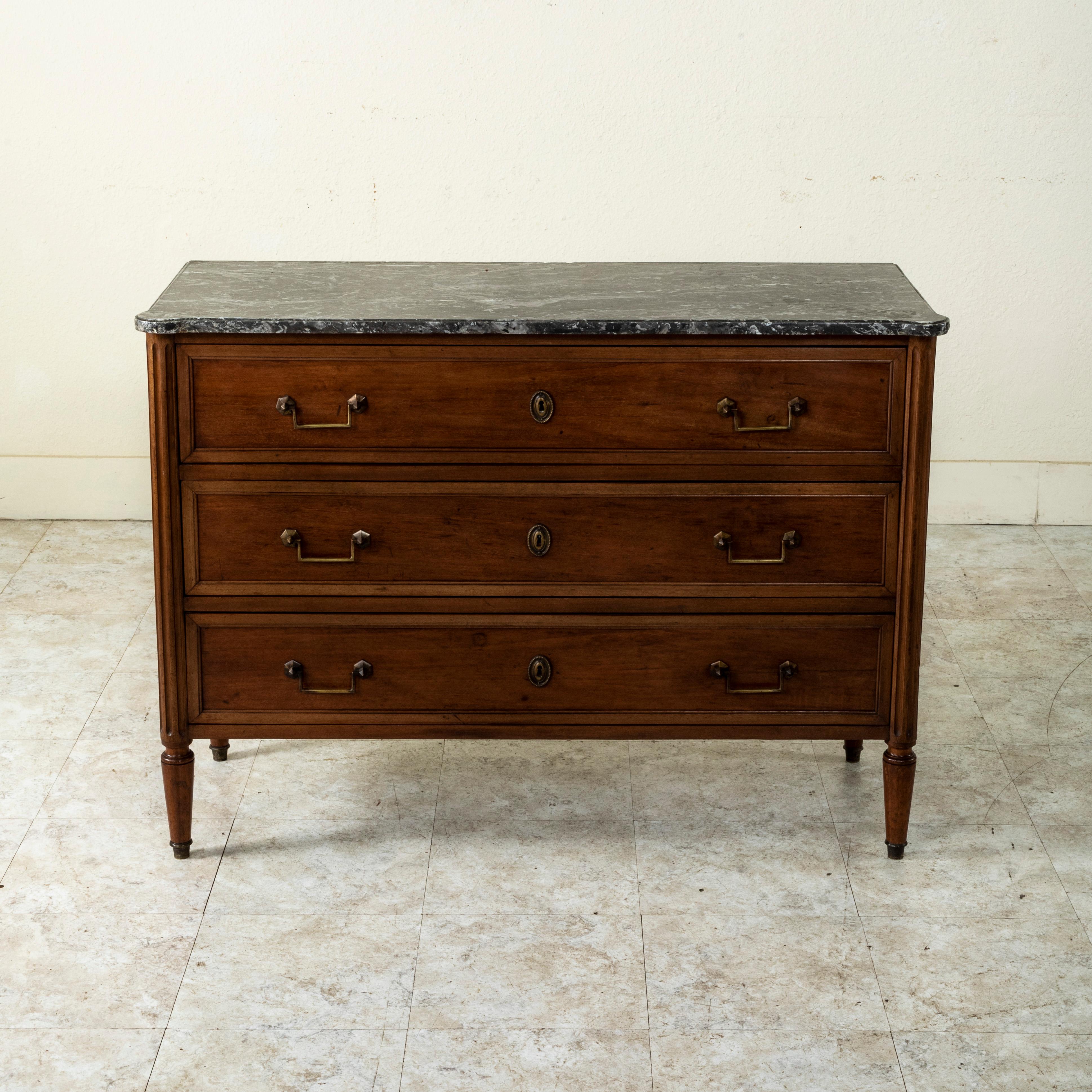 This early twentieth century French Louis XVI style mahogany commode or chest of drawers features a beveled Saint Anne marble top and fluted corners. Its three drawers of dovetail construction are fitted with classic beaded escutcheons and squared