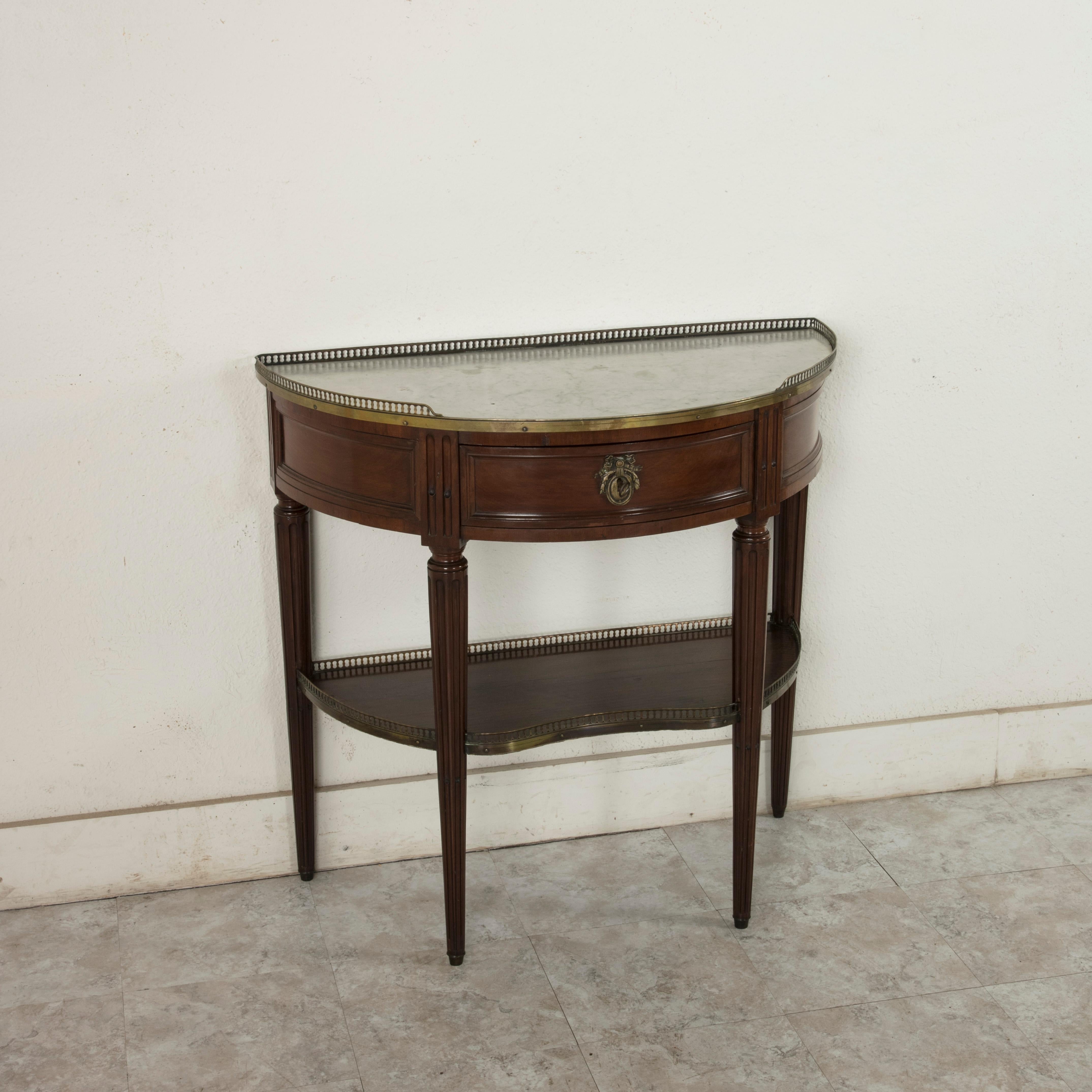 This French Louis XVI style mahogany demilune console table from the turn of the 20th century features a white marble top surrounded by a pierced bronze gallery. A single drawer of dovetail construction fits into the apron below and is finished with