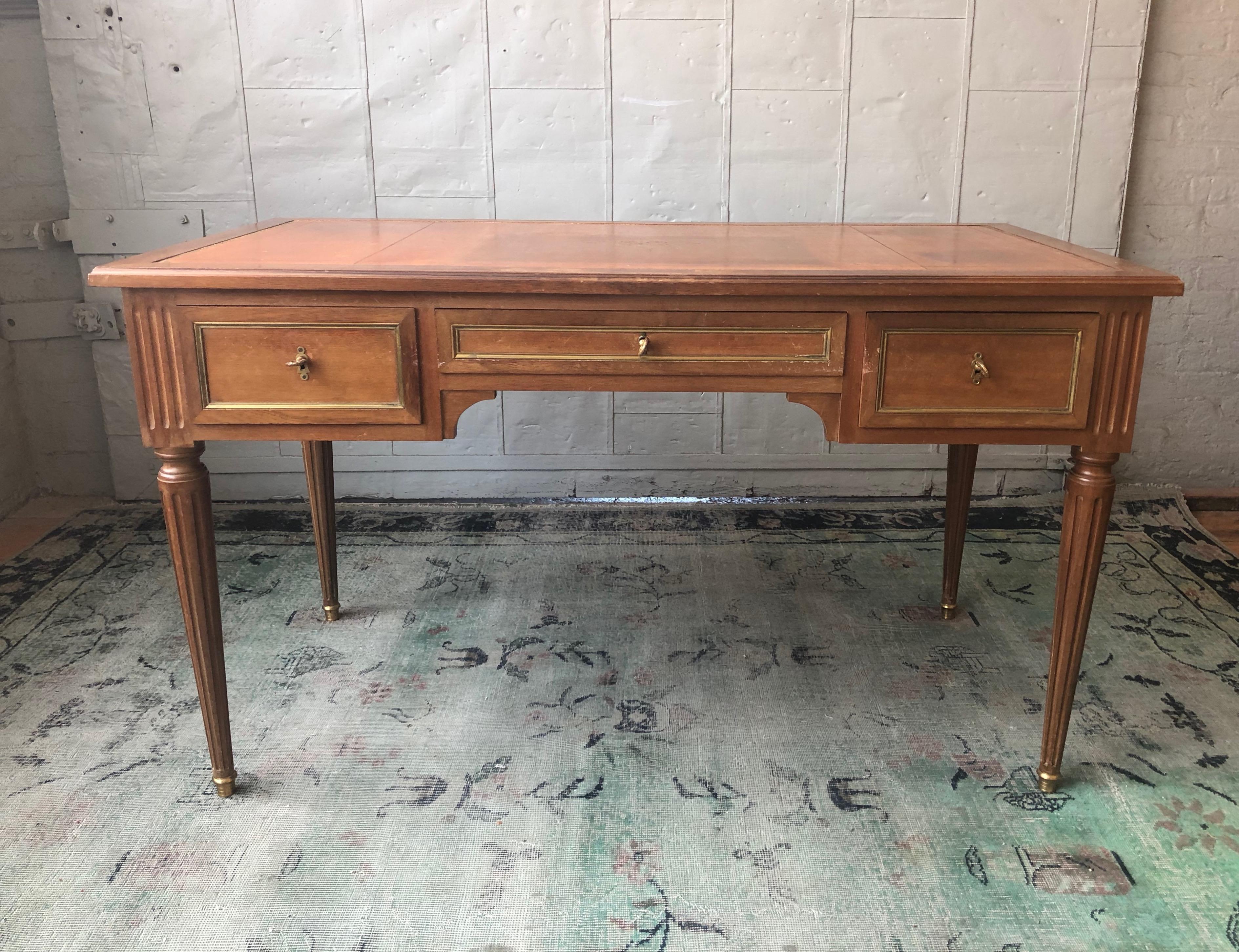 An elegant writing desk made in solid mahogany with brass trims and a tooled leather writing surface. The fluted and tapered legs are mounted on brass sabots. The three drawers each have functioning locks and keys. The backside of the desk is