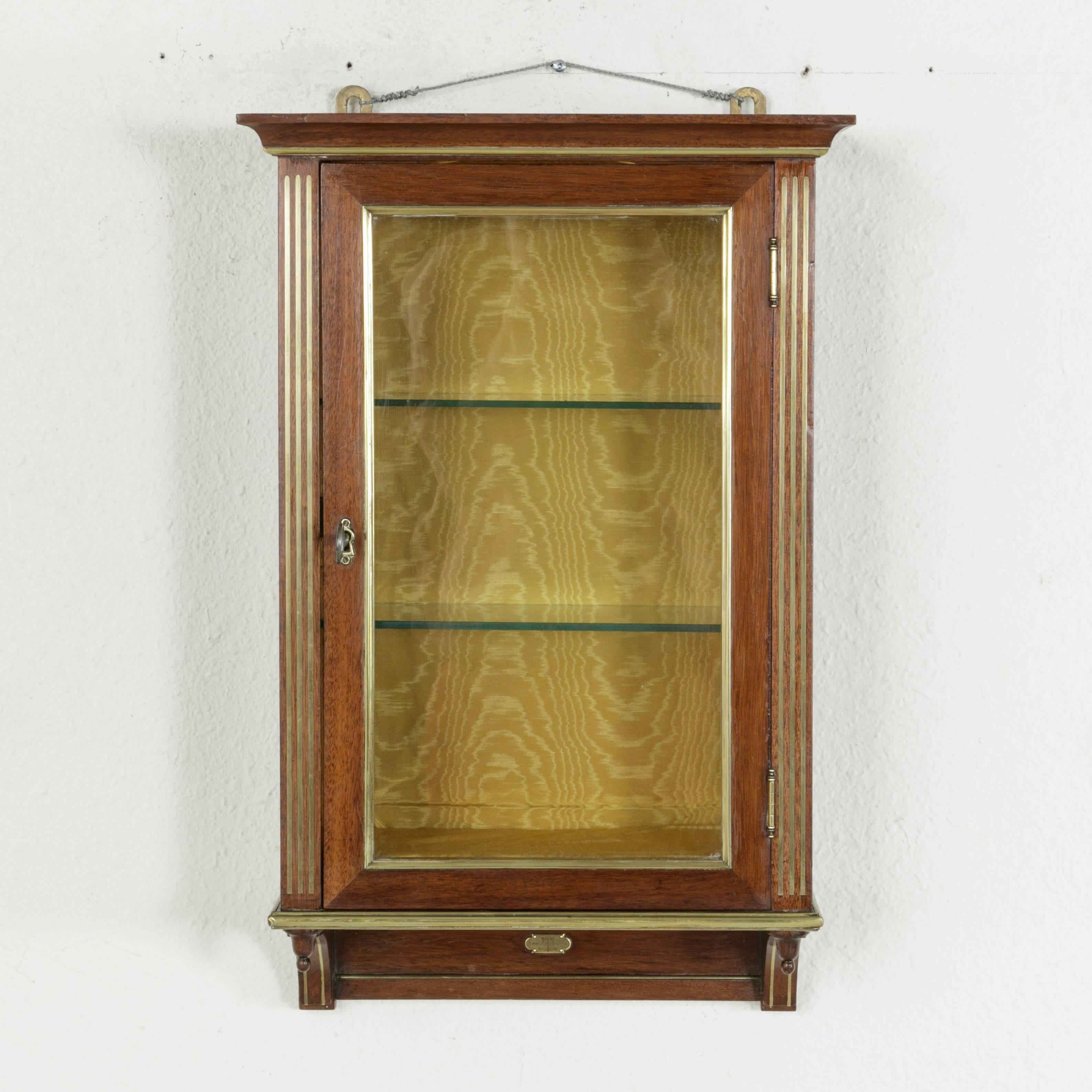 This early 20th century French Louis XVI style mahogany wall vitrine or display cabinet features fluted corners of bronze inlay as well as additional bronze detailing that accentuates its clean lines. The single door is framed in bronze and boasts