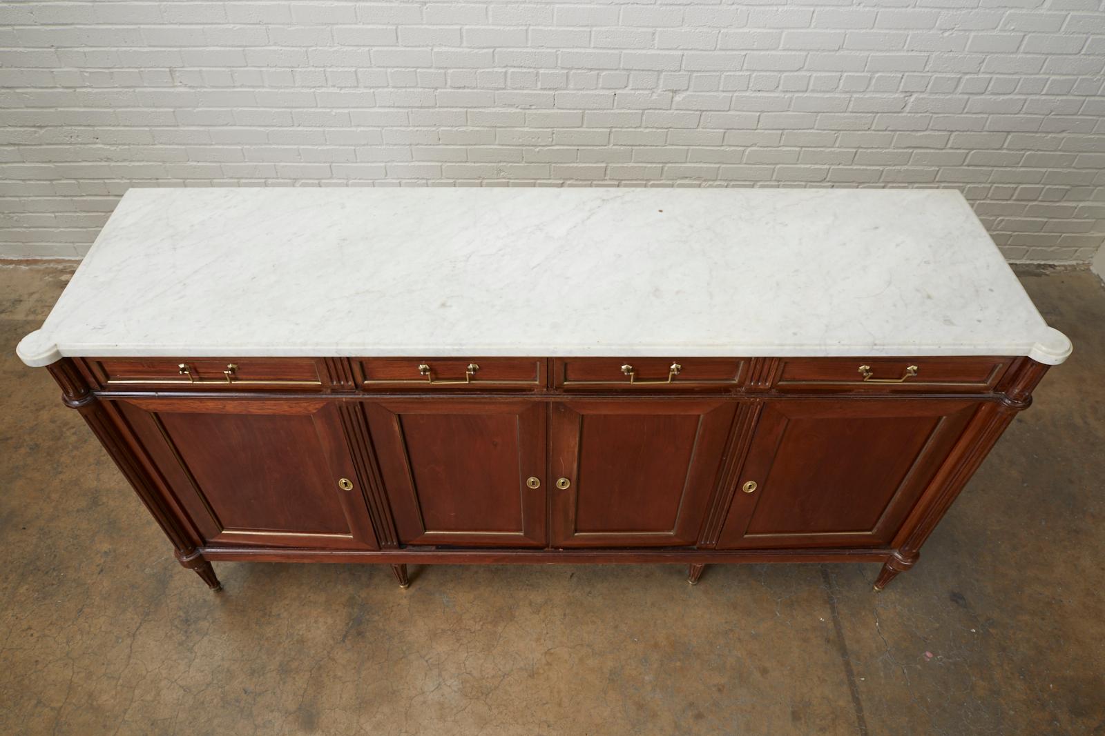 Distinctive French mahogany sideboard or buffet featuring a dramatic white Carrara marble top. Made in the grand Louis XVI Directoire style with fluted pilaster columns on each front corner. There are four storage drawers and four enfilade style