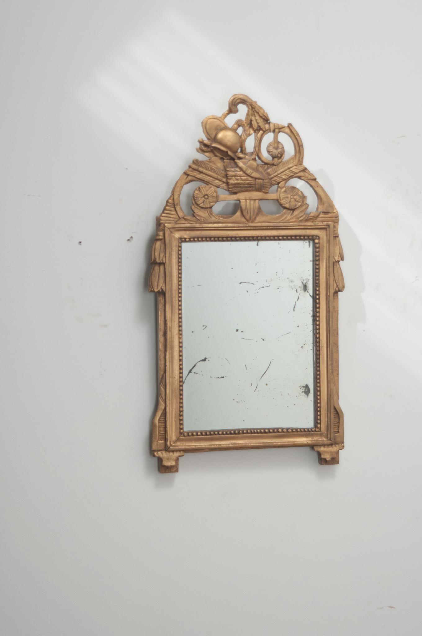 A French petite mirror is the Louis XVI style. The painted gilt carved frame has a pierced crest depicting a military helmet, sword, shield, quivers and ax. The original mirror plate is surrounded by the mirror frame’s beaded trim. Some repairs have