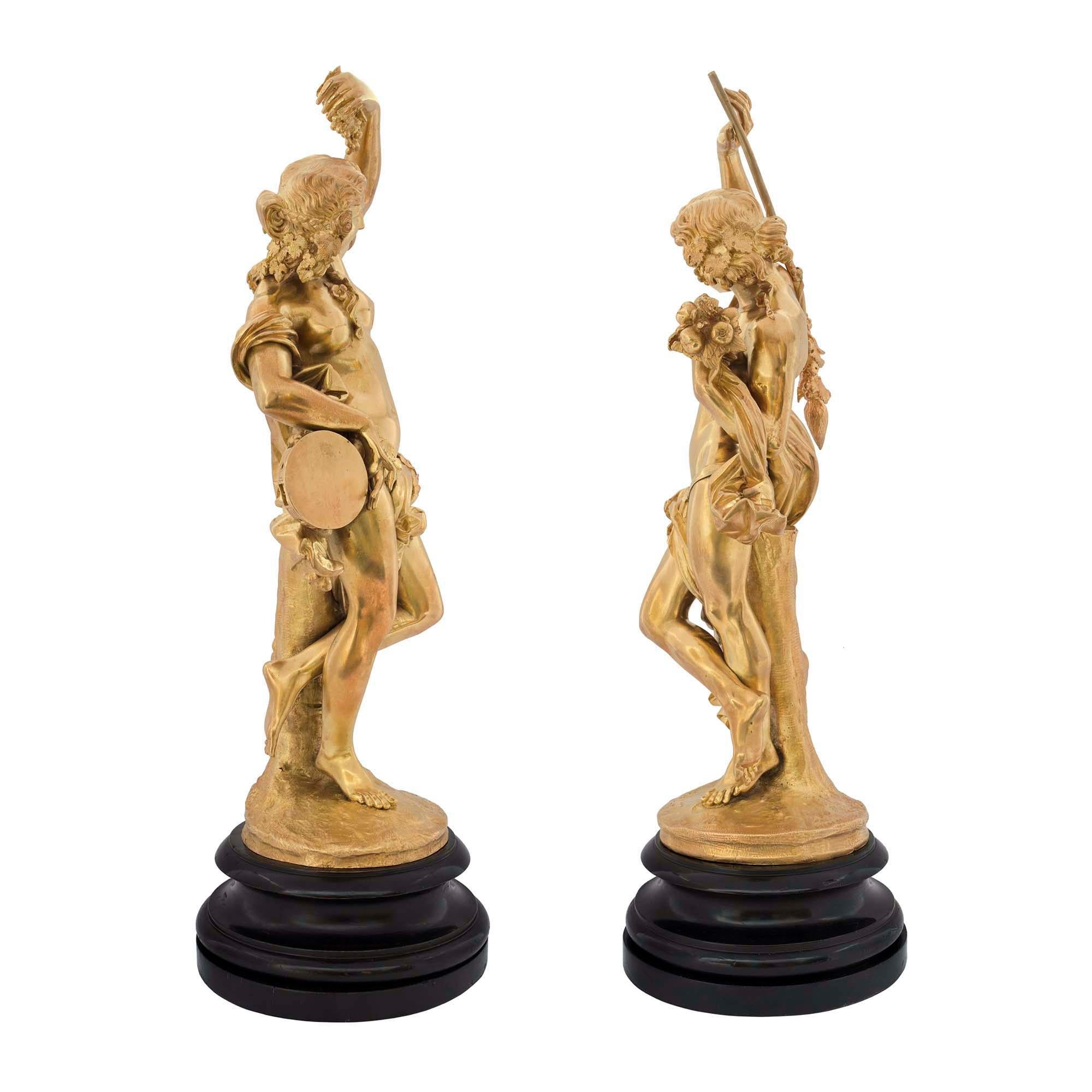 Paire de statues de fête en bronze doré de style Louis XVI, signée Devaulx. À gauche, elle est appuyée sur une souche d'arbre, drapée dans un vêtement fluide, avec un tambourin dans une main et une grappe de raisin dans l'autre. Le mâle à droite est