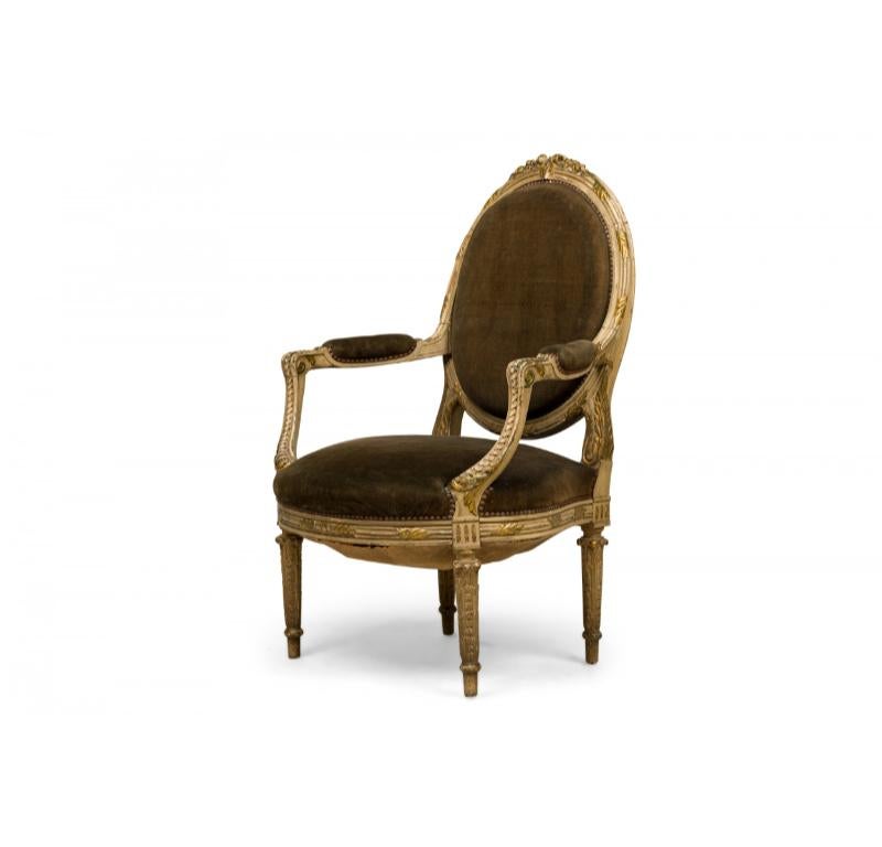 French Louis XVI-style (19/20th Century) armchair having green velvet upholstered oval backs, nailhead trim, and frames with distressed grey painted finish and gilt trim with carved leafed pattern topped by rose crests, standing on tapered legs.