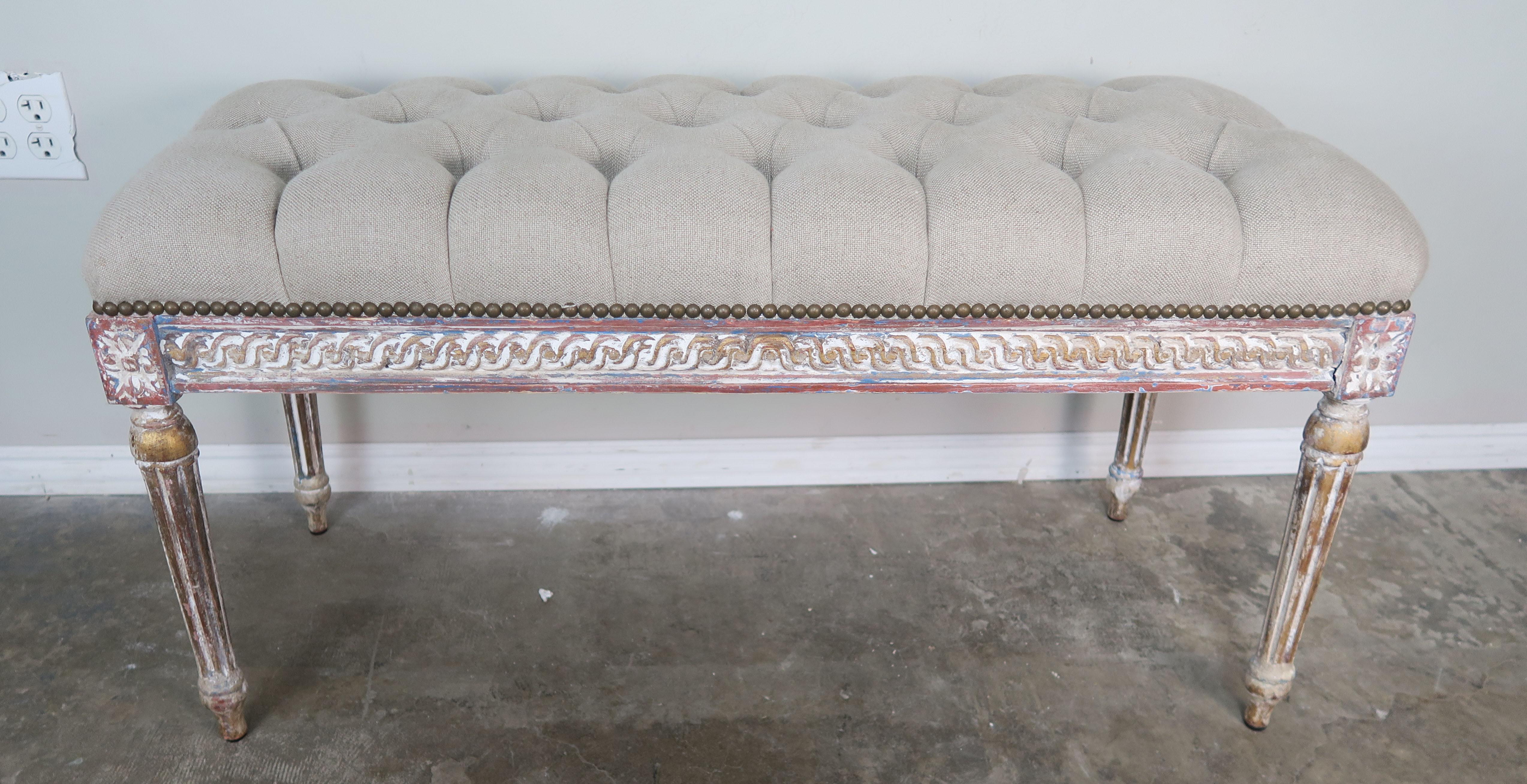 French Louis XVI style painted bench in muted shades of rust, blue, antique white, and gold leaf accents. The bench stands of four straight fluted legs and is newly upholstered in a washed tufted Belgium linen with antique brass nailhead trim detail.