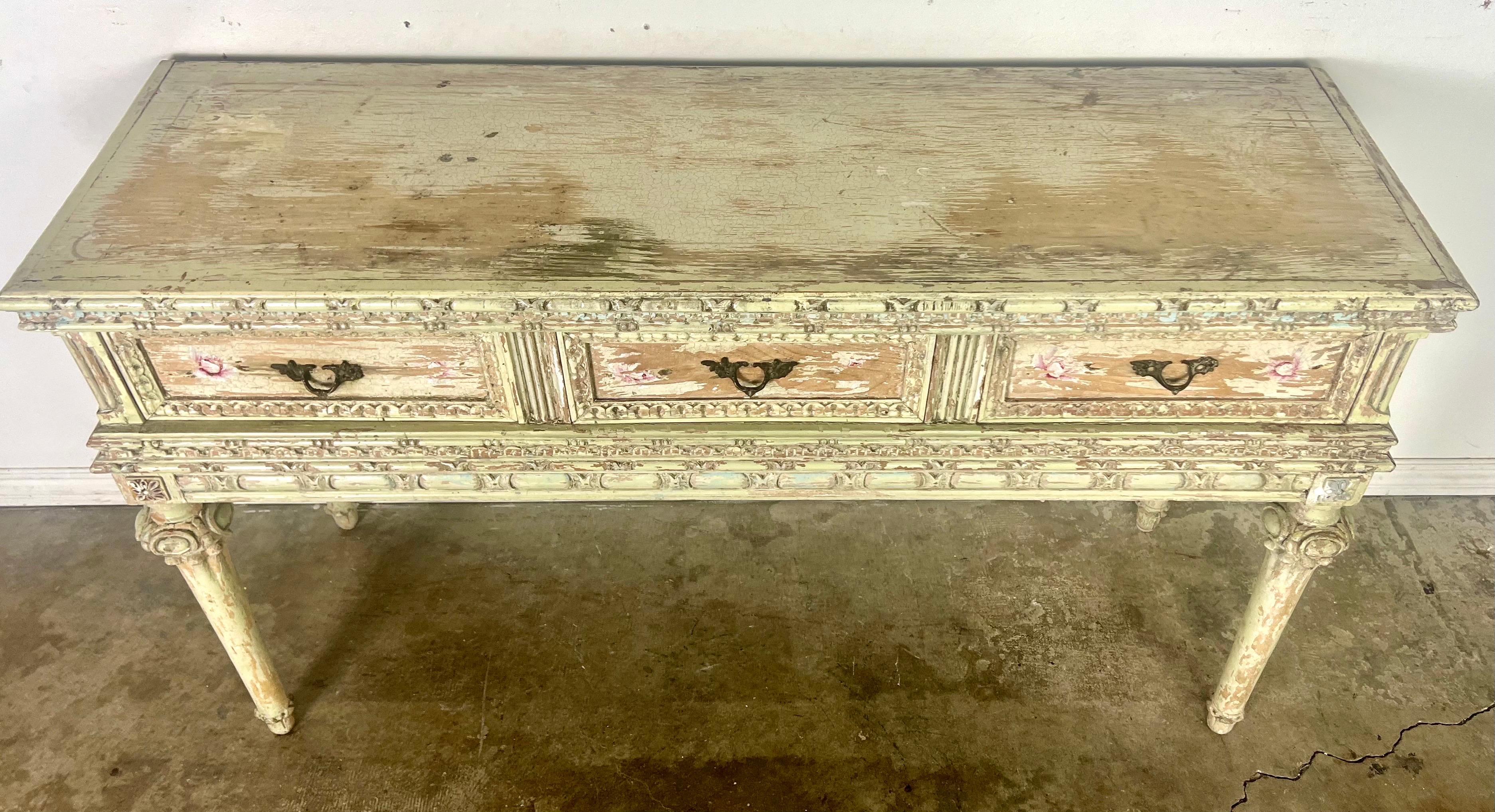 Louis XVI style French painted console table with decorative flourishes.  It features a distressed finish, classical motifs, and fluted legs inspired by neoclassical design elements.  The ornate carving and painted details add to its charm. Remnants