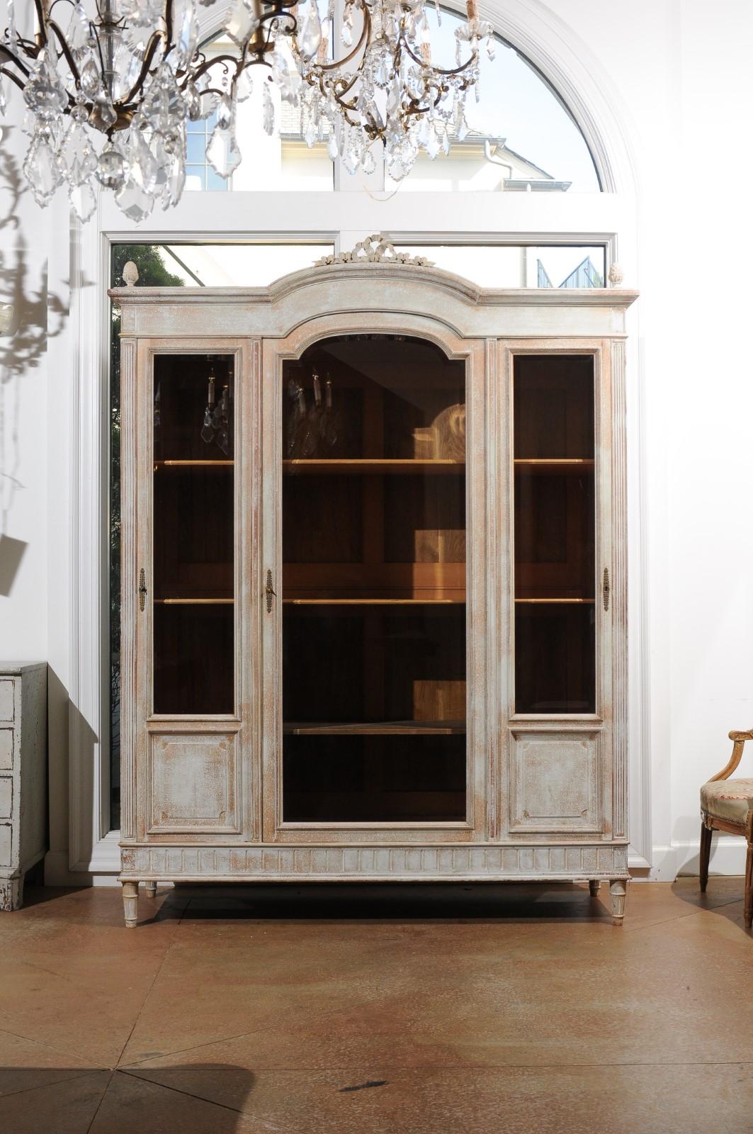 A French Louis XVI style painted wood cabinet from the 20th century with glass doors, distressed finish, 'chapeau de gendarme' cornice and carved ribbon crest. Born in France during the 20th century, this exquisite painted wood cabinet presents the