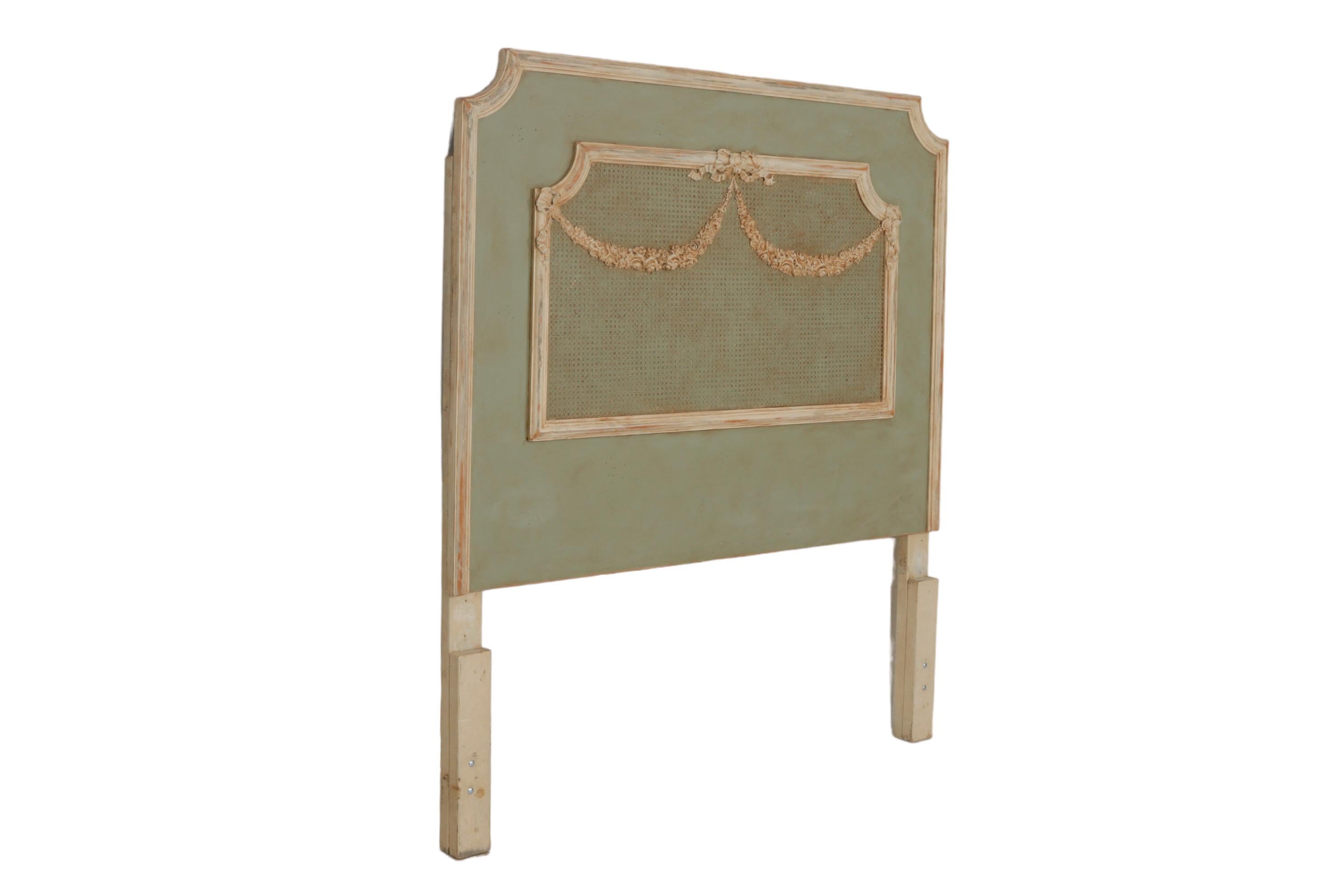 A French Louis XVI style queen size headboard. A caned center is decorated with carved ribbon and rose swag details. Painted in a sage green and trimmed with cream, with subtle distressed edges and an antique patina. Made by Gildid of Atlanta.