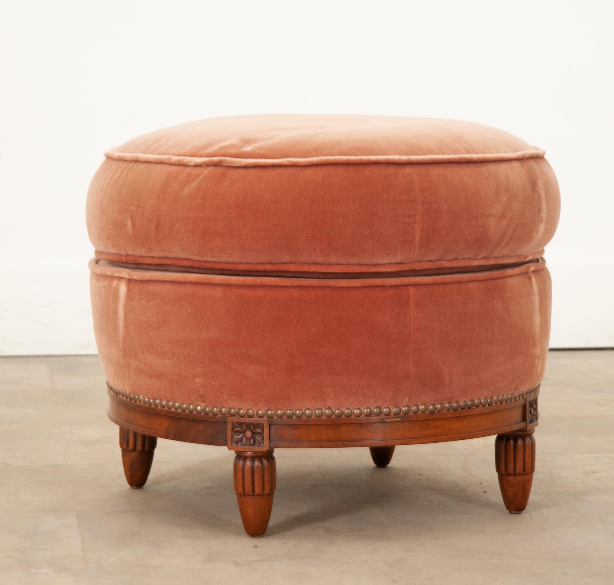 A pretty and plush pouf, ottoman, or footstool in the Louis XVI style with elegant hand-carved elements and coral pink velvet upholstery. Its round solid mahogany wood frame is beautiful with intricately carved framed rosettes on each of its four